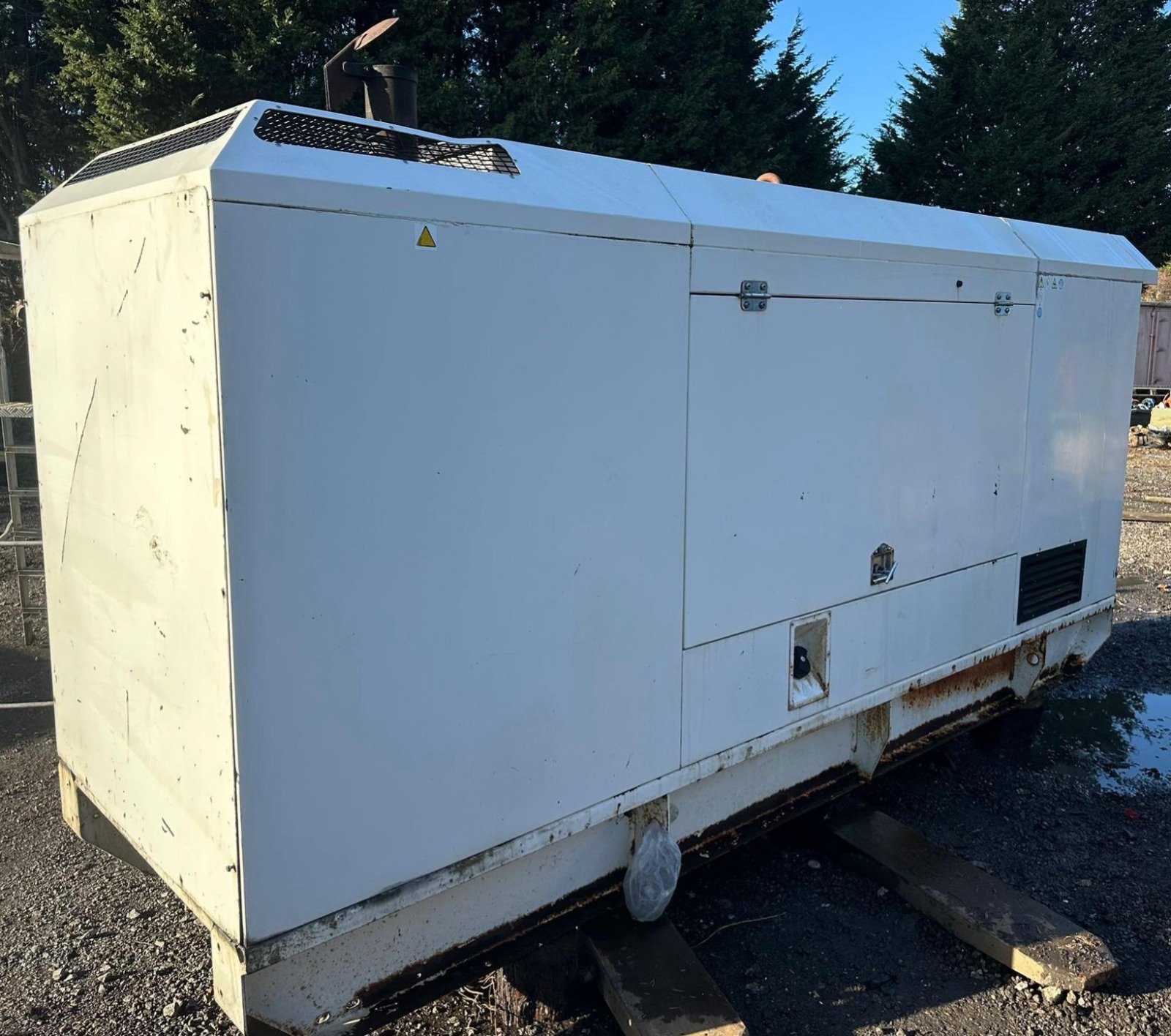 A large white industrial generator or transformer with rust at the bottom, placed outdoors on wooden supports with gravel ground and trees in the background.
