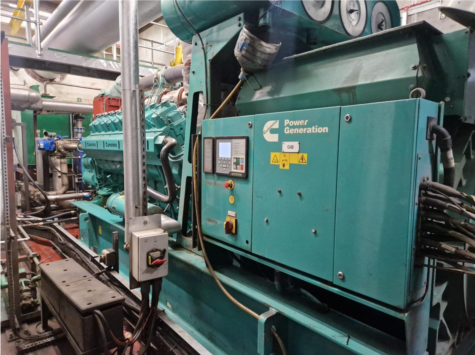 An industrial generator with a teal-colored exterior, labeled 'Power Generation' and 'Cummins', surrounded by pipes, gauges, and control panels.