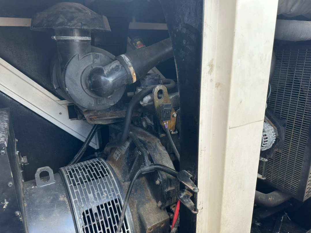 Close-up of a mechanical engine part with a black pipe, wiring, and metal components, some showing rust, in an industrial or automotive setting.