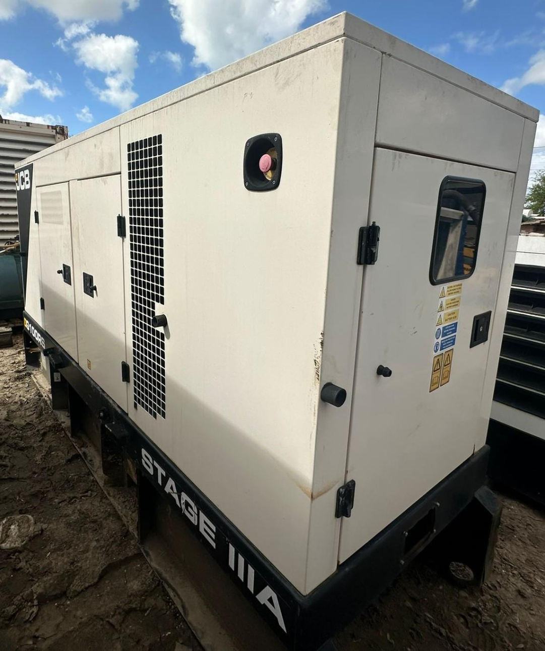A large portable industrial generator with white and black exterior, labeled 'STAGE II' and 'JCB', sitting on dirt ground outdoors with a partly cloudy sky.