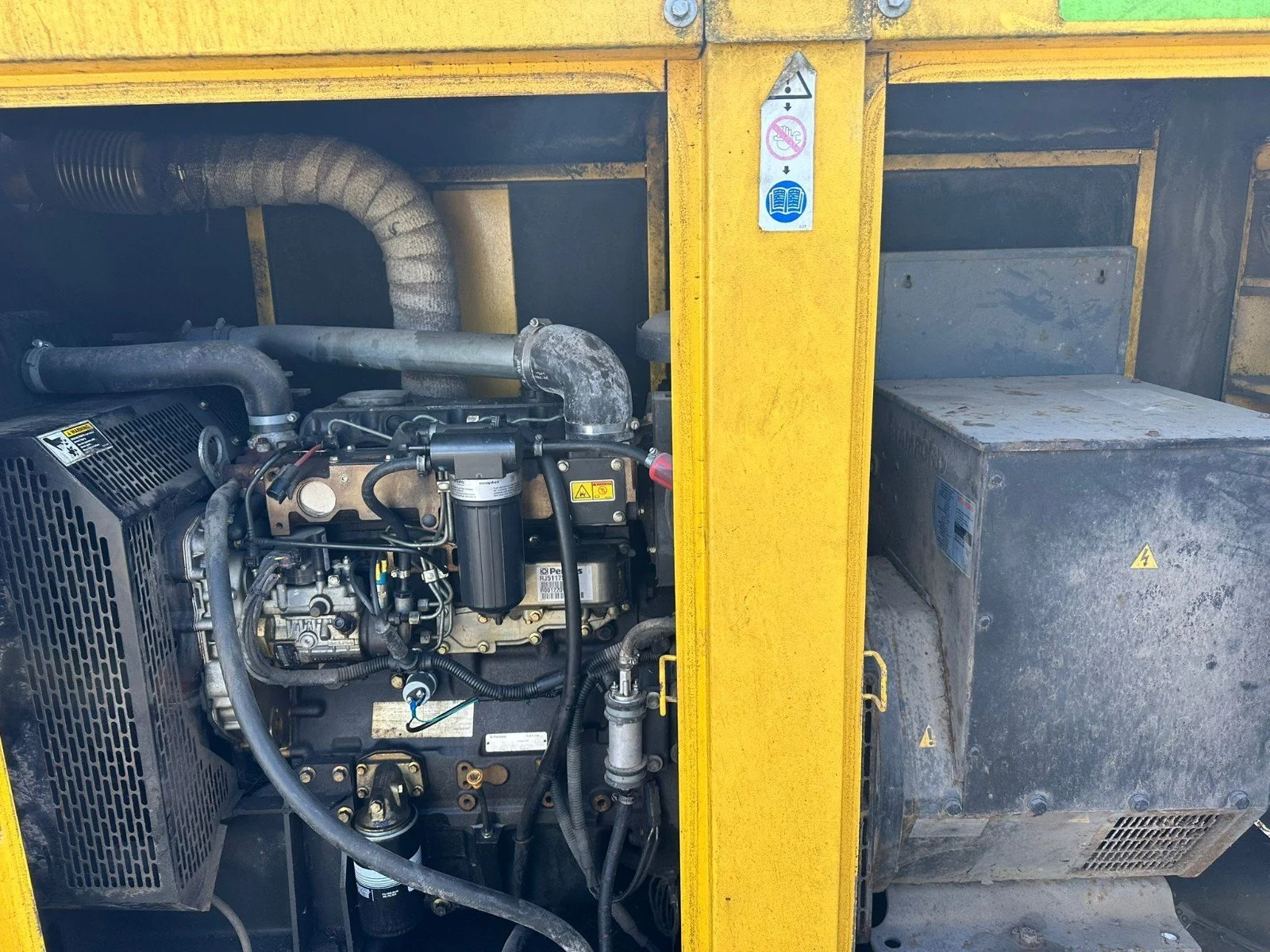 Close-up of an industrial generator engine with various pipes, filters, and metal components, partially enclosed within a yellow frame.
