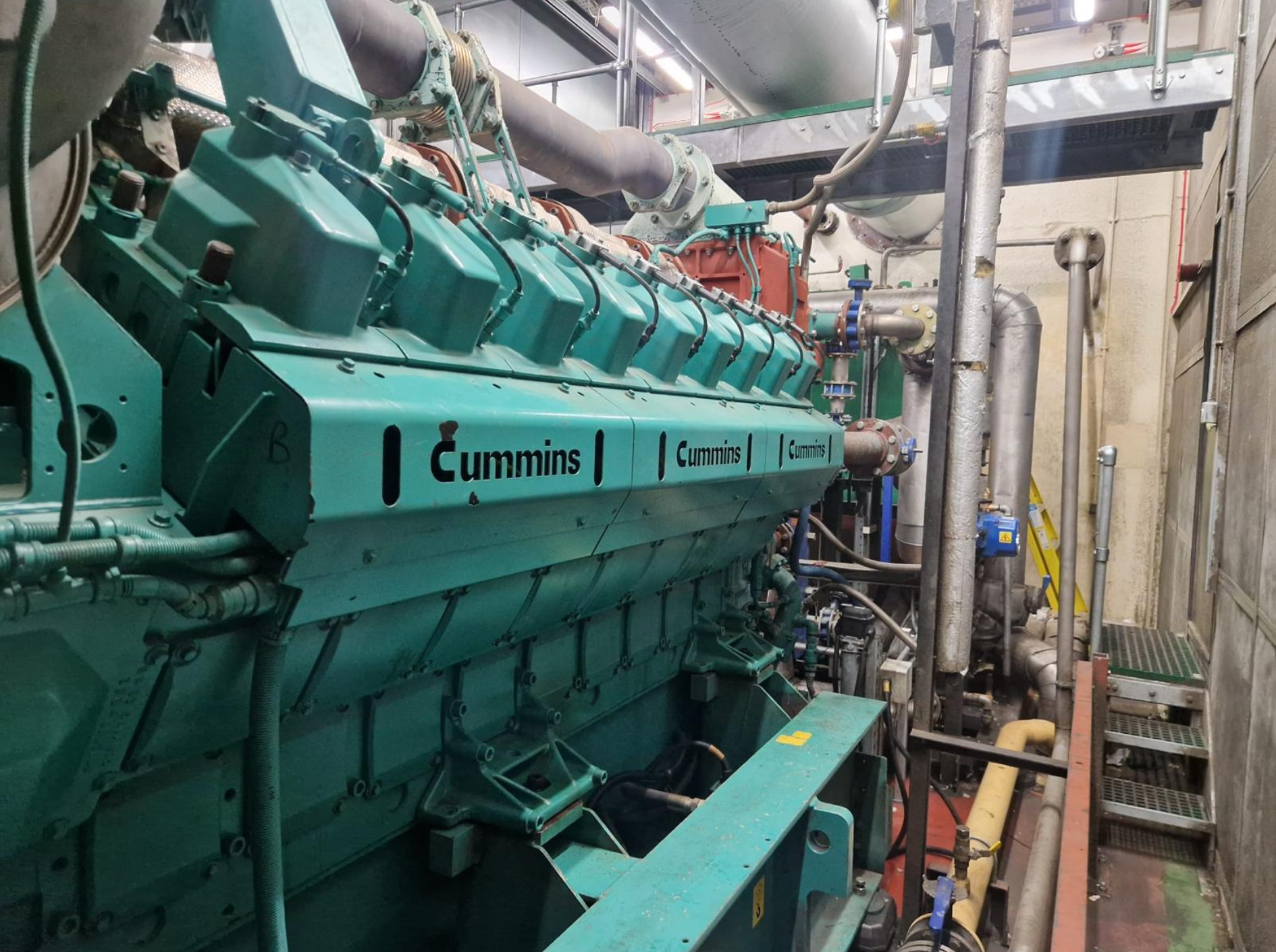Industrial engine or generator inside a mechanical room with pipes and support structures.