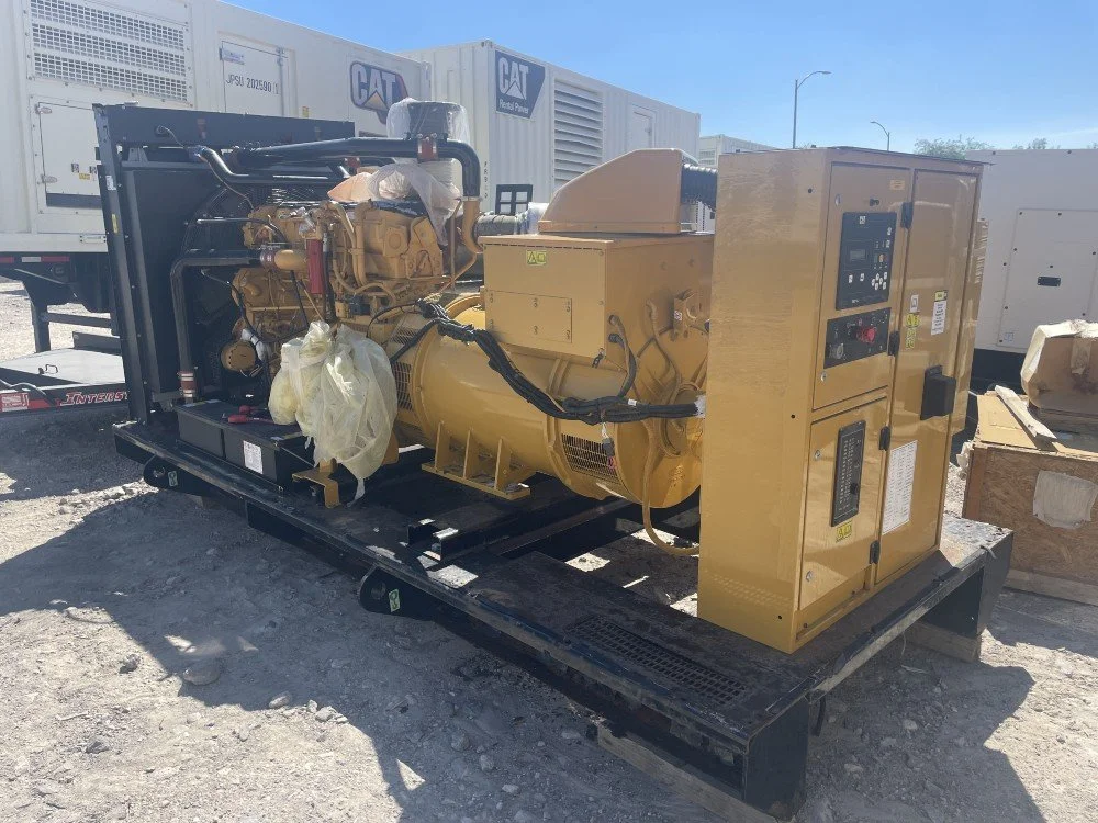 A large yellow industrial generator with control panel, mounted on a black metal frame outdoors, with trailers and equipment in the background.