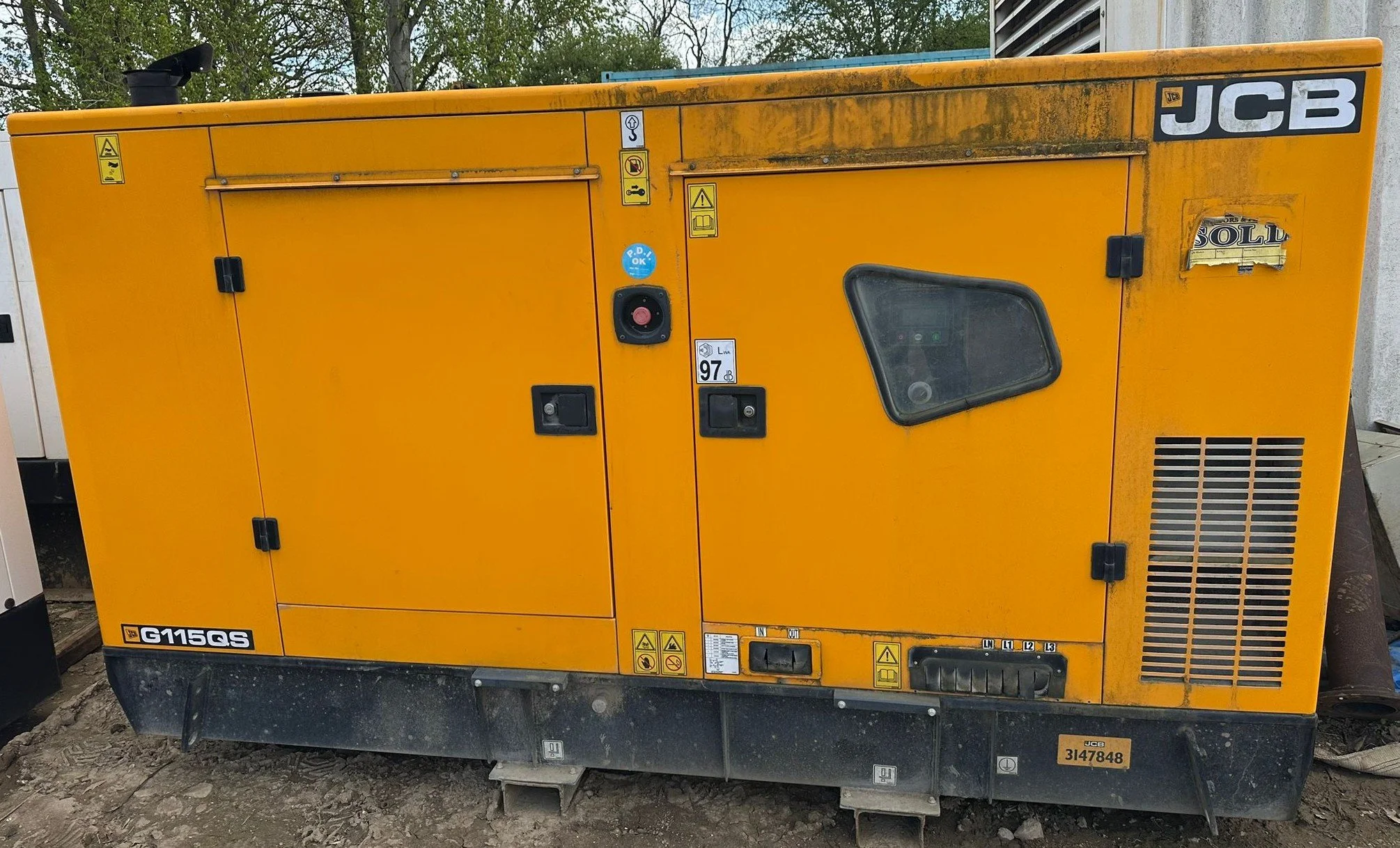 Yellow industrial generator with various warning labels, a small window, and a black exhaust pipe on top, placed outdoors on dirt ground.