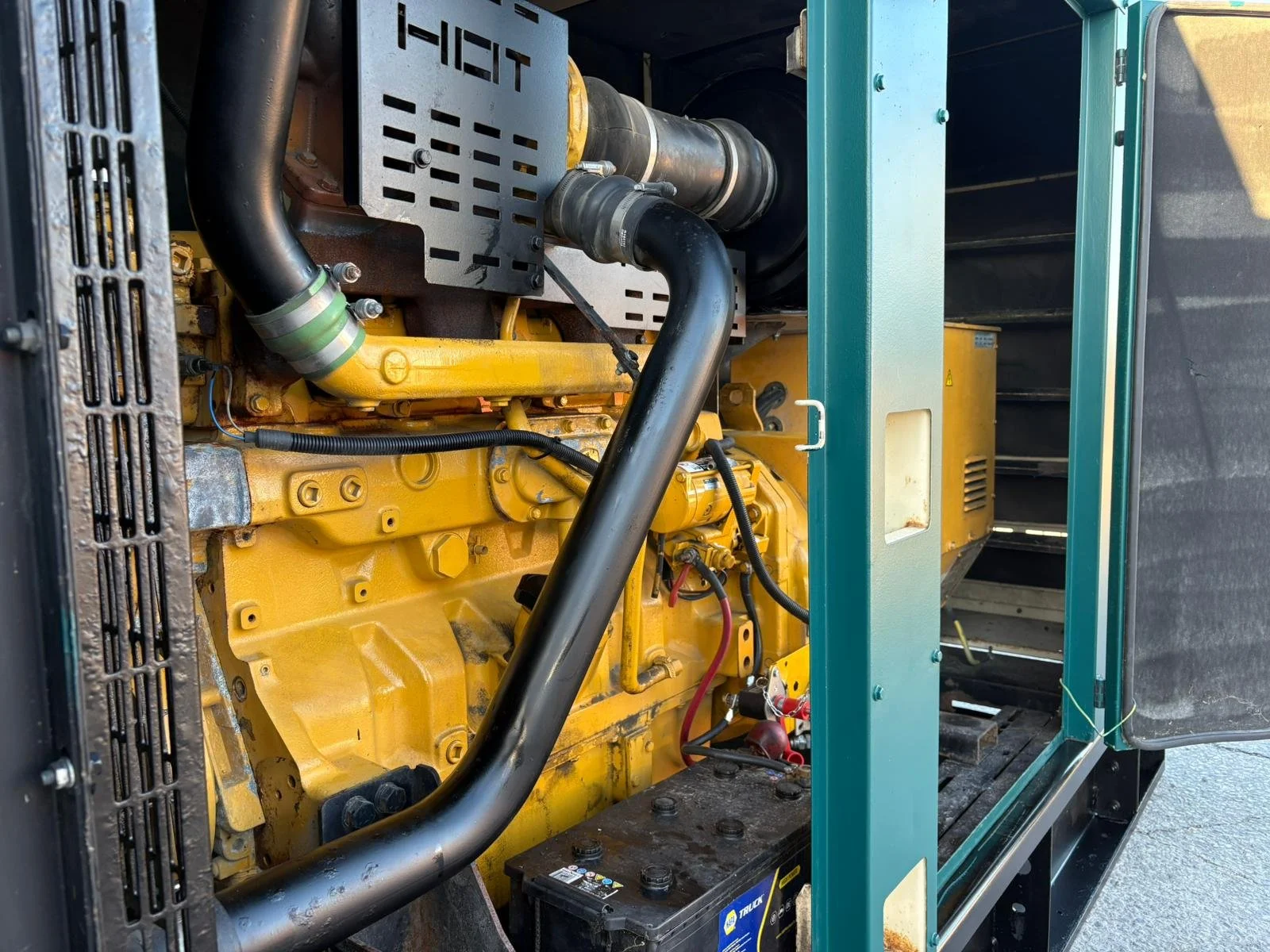 Close-up of a yellow industrial generator engine with black and green hoses and a black battery inside a metal frame.