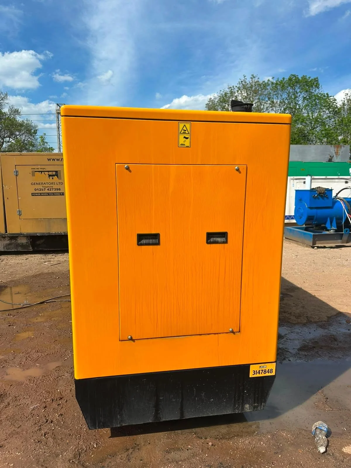 Orange industrial generator on dirt ground with a warning label and a black base, surrounded by other industrial equipment under a blue sky with some clouds.