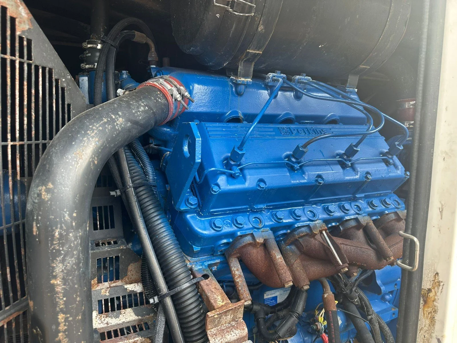 Close-up view of a blue Perkins engine with visible hoses, wires, and rusty exhaust manifold in an industrial setting.