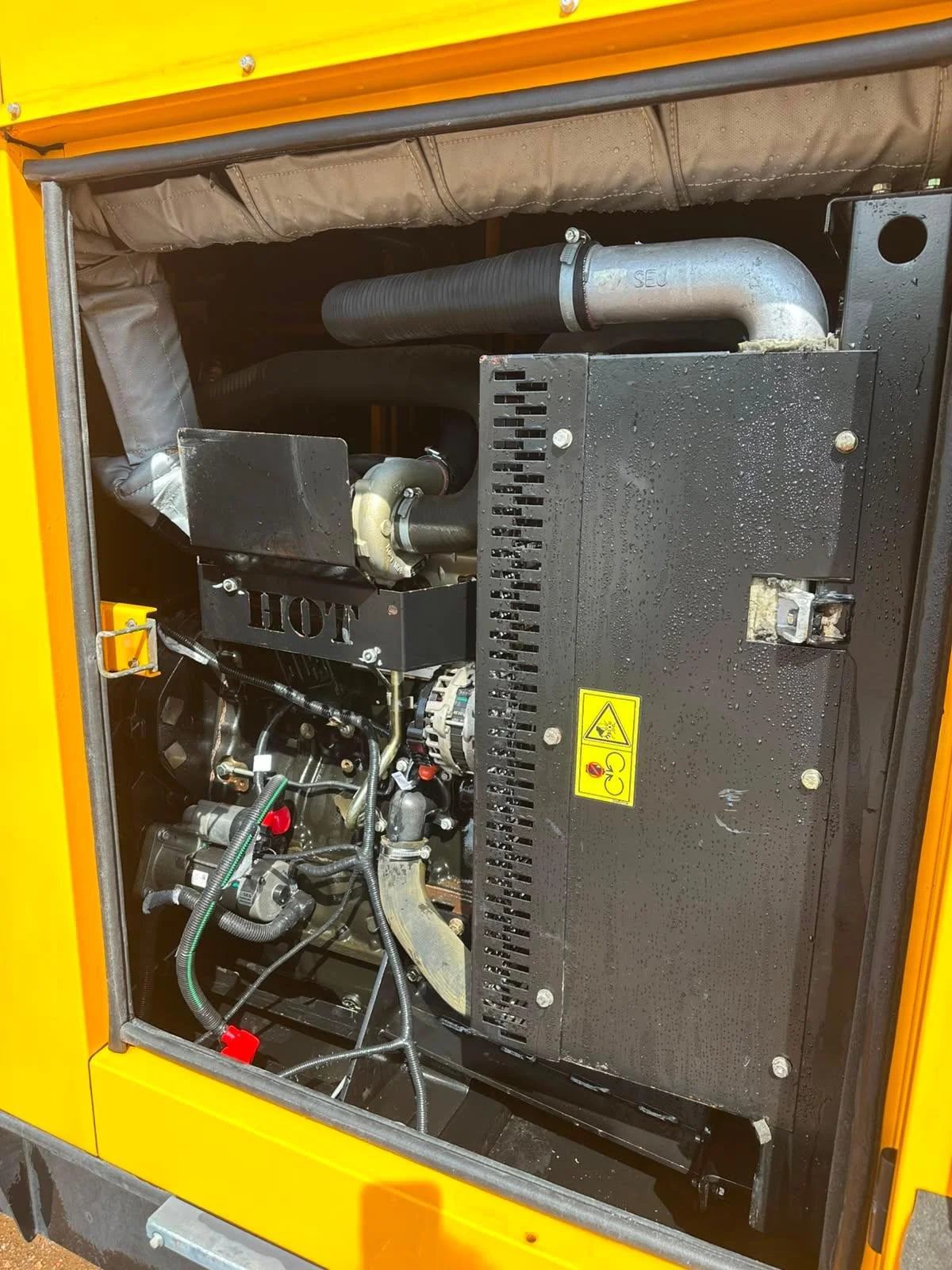 The image shows the internal components of a generator, including pipes, wires, and a black metal box with a yellow warning label.