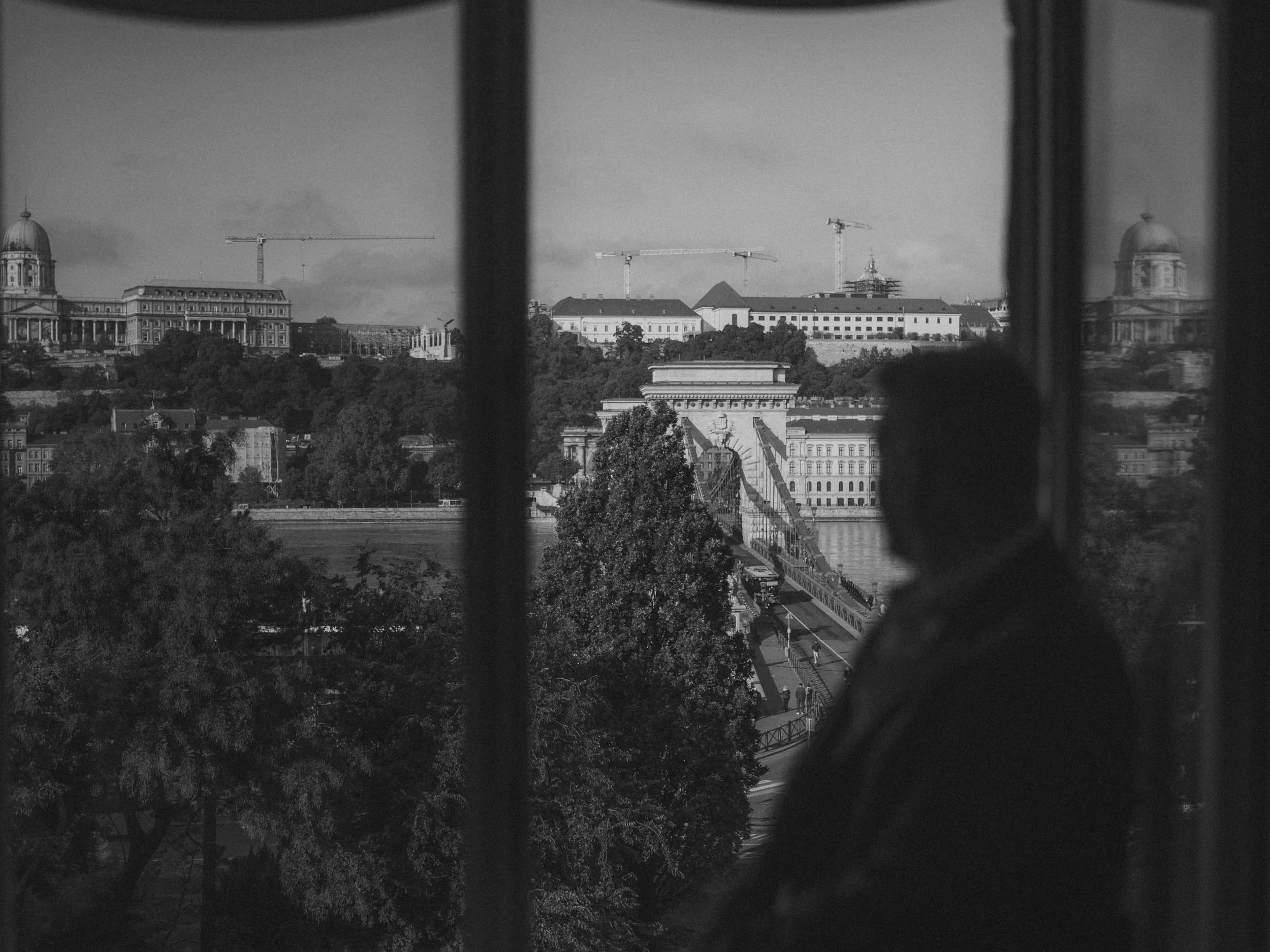 Black and white photo of a person looking out a window at a cityscape with historic buildings and cranes in the background, bridge over a river in the middle ground.