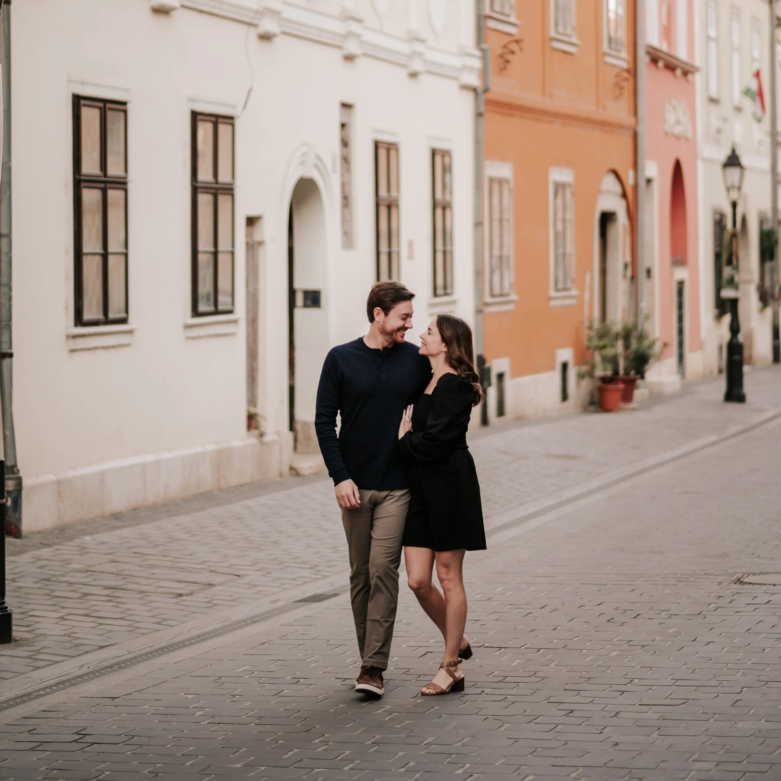 A couple walking arm in arm on a cobblestone street, smiling at each other, with European-style buildings in the background.