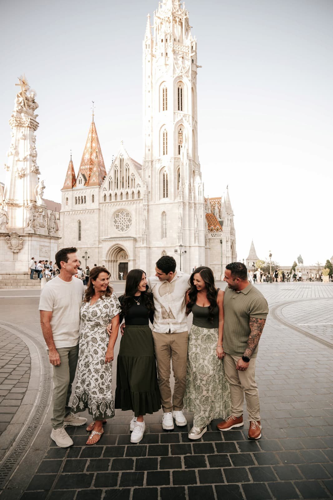 Group of six people standing together in front of a large, ornate church with a tall spire and a statue on the side, smiling and enjoying each other's company.