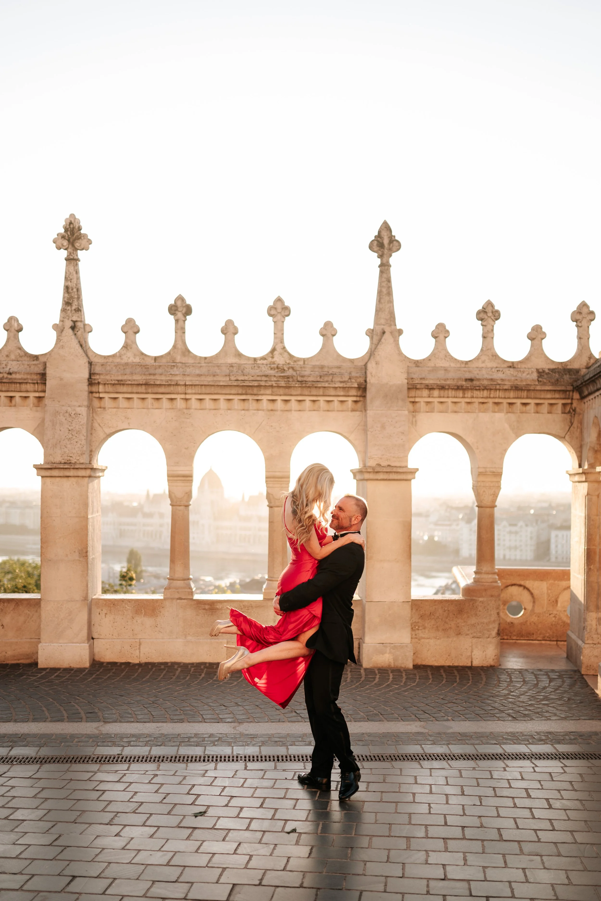 He lifts her off her feet in front of the sunlit arcade at Fisherman’s Bastion