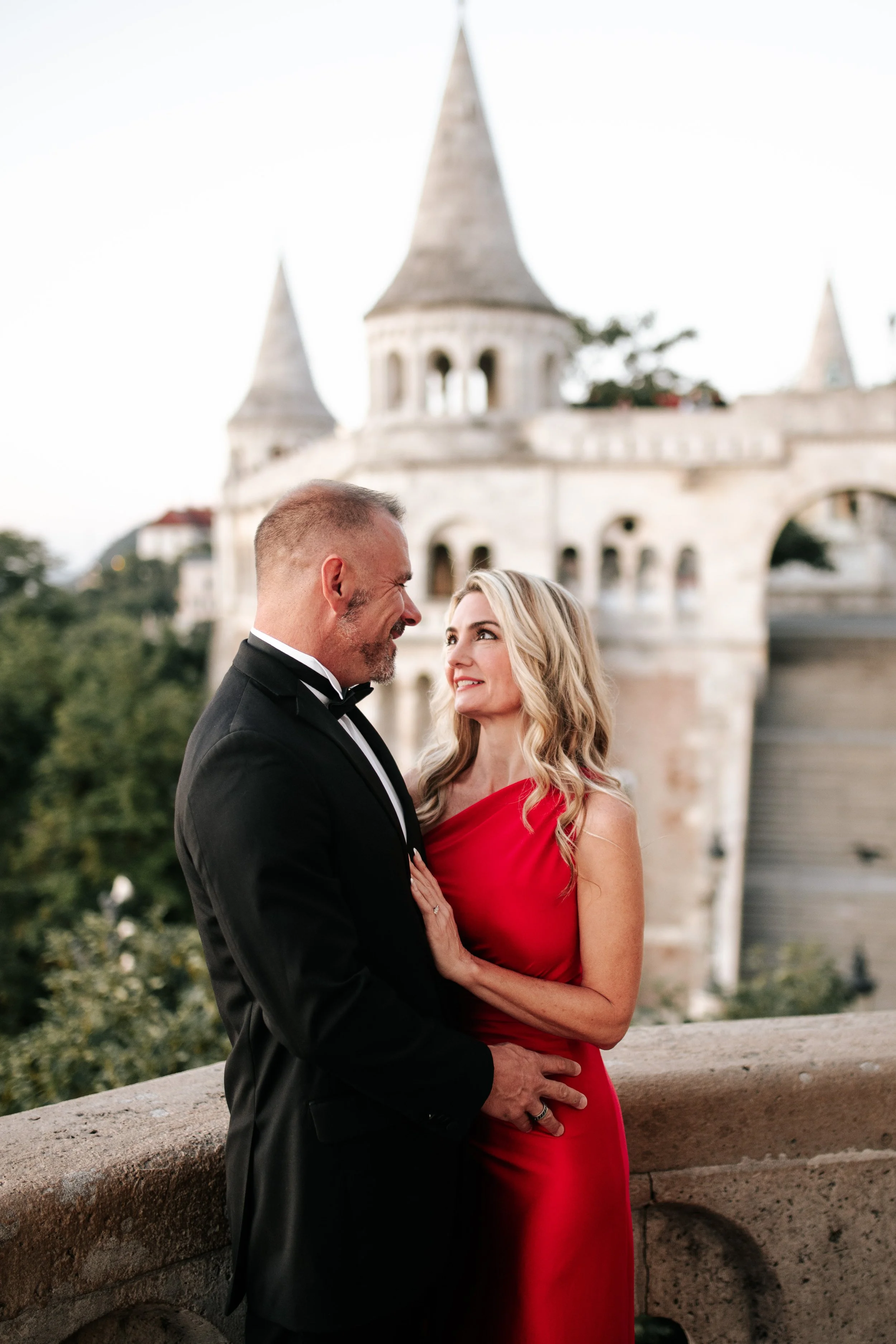 Smiling at each other beside the balustrade, her red dress against the pale stone towers