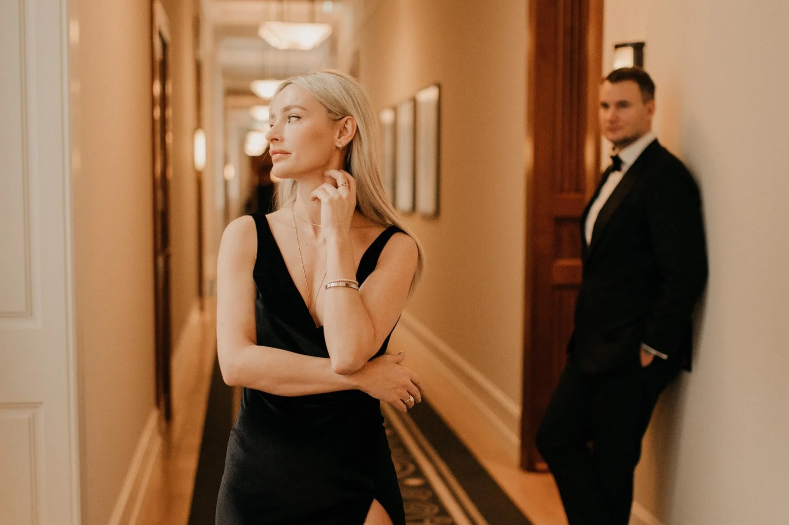 A woman in a black evening gown standing in a hallway, looking to the side with a contemplative expression, while a man in a tuxedo leans against the wall in the background.