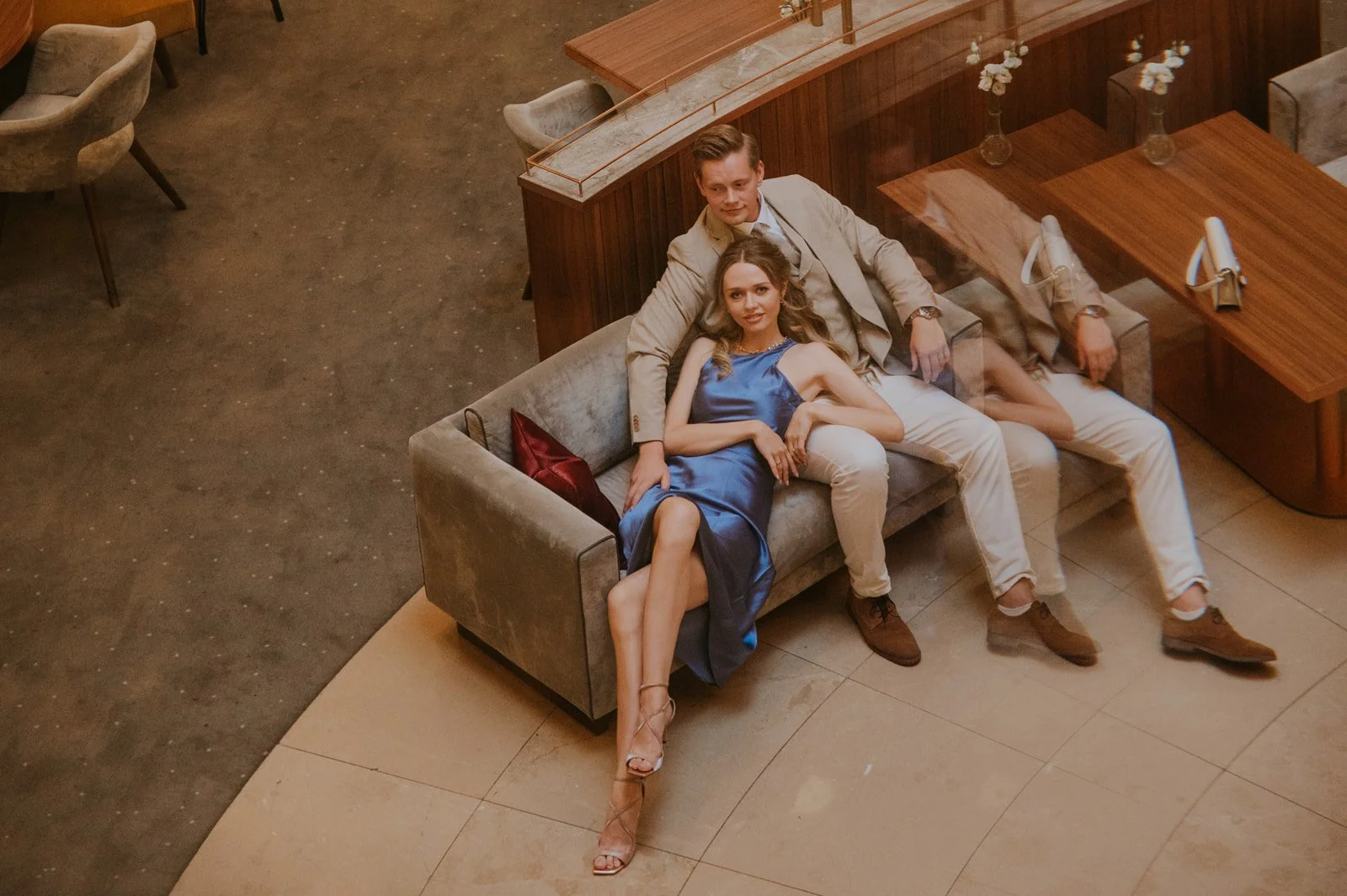 A man and a woman are sitting on a gray couch in a stylish indoor space, with the woman wearing a blue dress and the man in a beige suit jacket and white pants. The woman is lying back with her legs crossed, and the man is leaning back behind her, with their arms around each other.