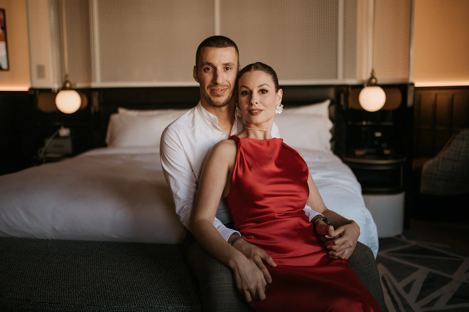 A couple sitting together on a chair in a hotel room, with the bed behind them. The woman is wearing a red dress and jewelry. The man is wearing a white shirt.
