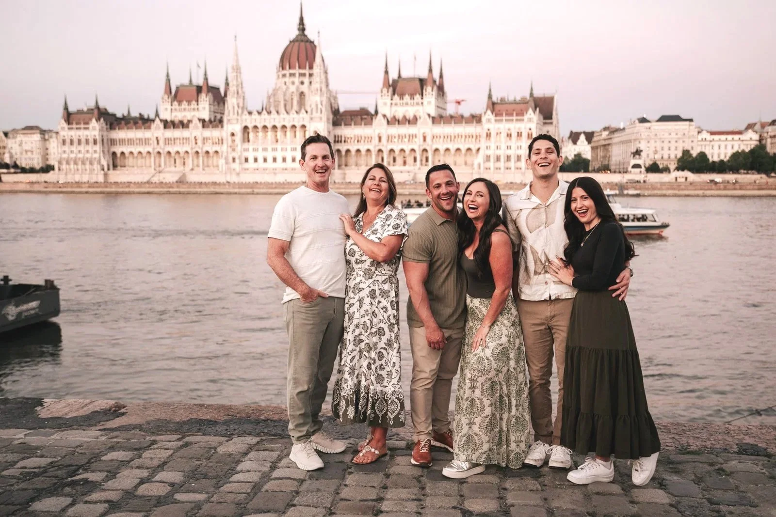 A group of six friends standing on a cobblestone walkway by a river, smiling, with the Hungarian Parliament Building in Budapest visible in the background.