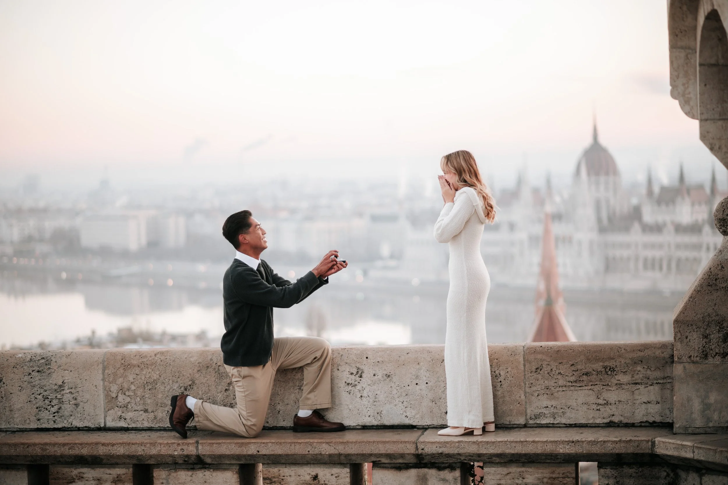 A man proposing marriage to a woman on a historic building balcony with a cityscape background at sunset.