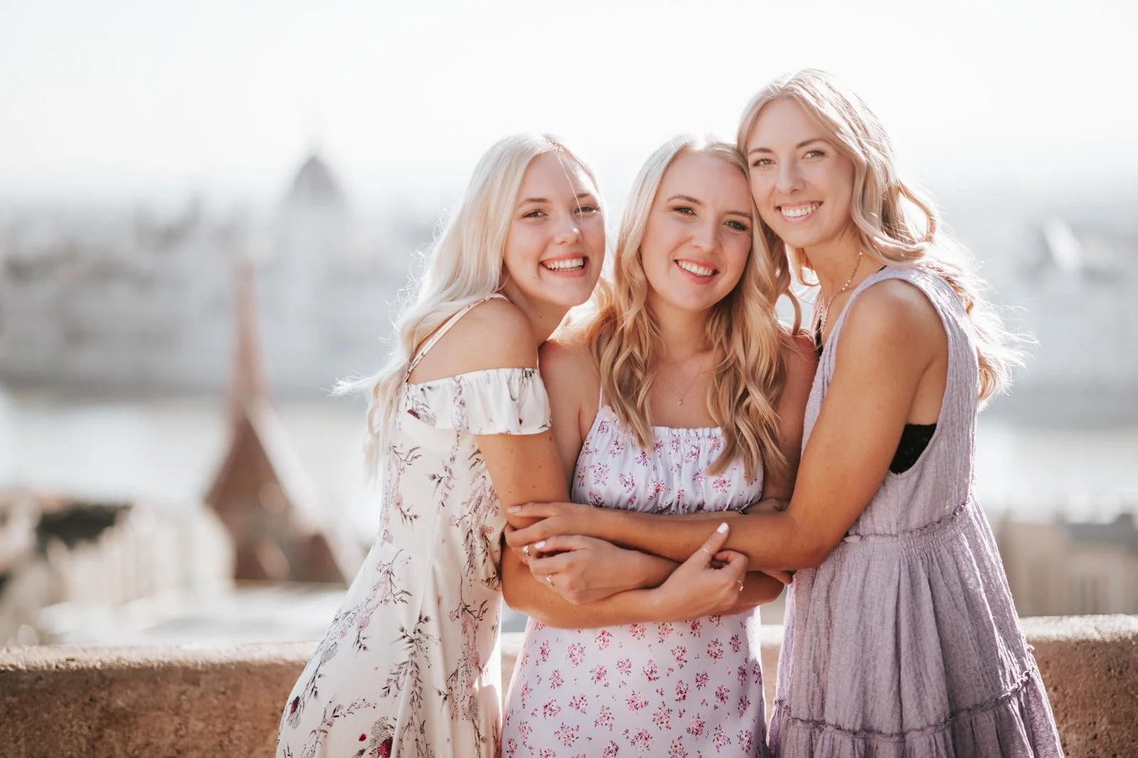 Three young women with blonde hair smiling and hugging outdoors with a cityscape and water in the background.