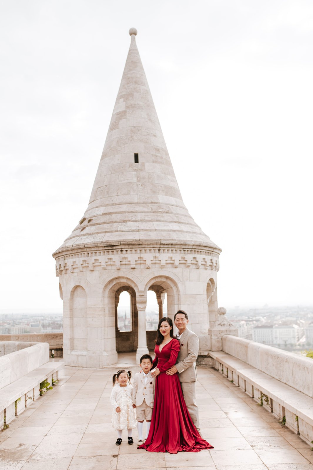 A family of four standing on a rooftop with a historic white stone tower in the background, city skyline visible in the distance, dressed in semi-formal attire.