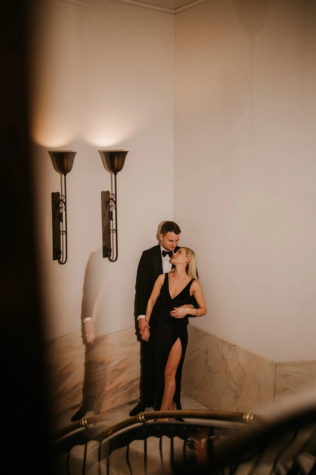 A man in a tuxedo and a woman in a black evening gown are standing close together in a corner of a room, holding hands and looking at each other affectionately, with wall sconces on the wall behind them.