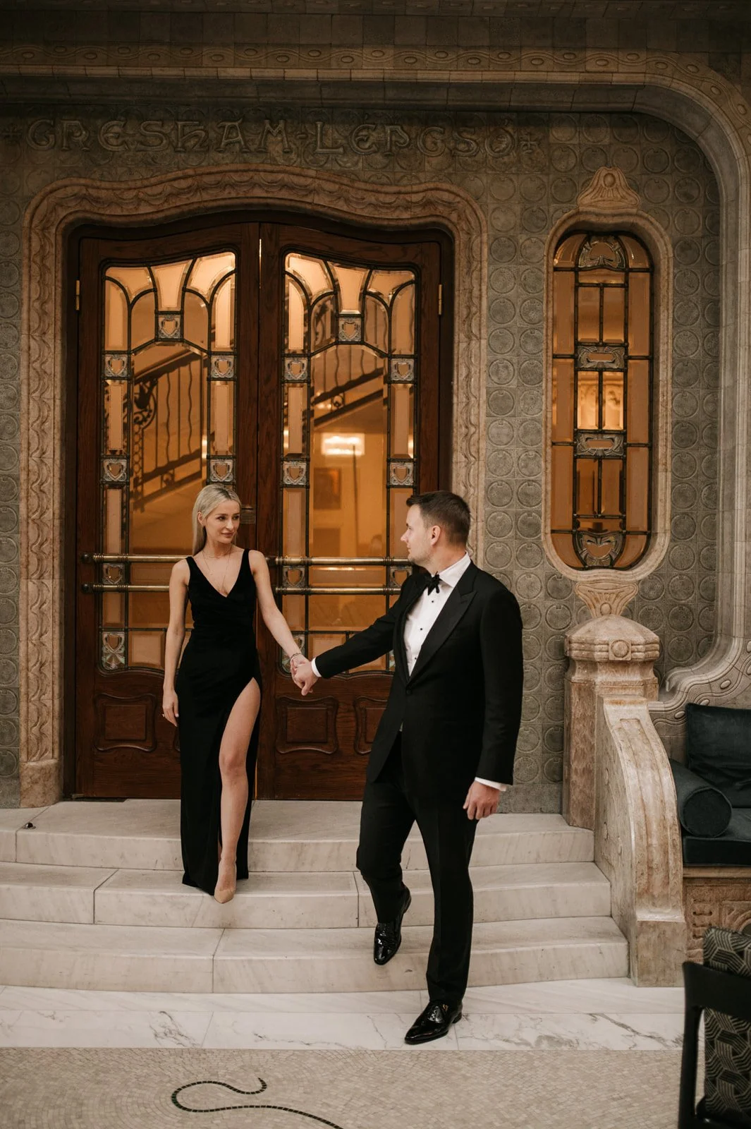 A couple dressed in formal evening wear standing on stairs in front of ornate wooden door; woman in black gown with high slit, man in tuxedo, holding hands.