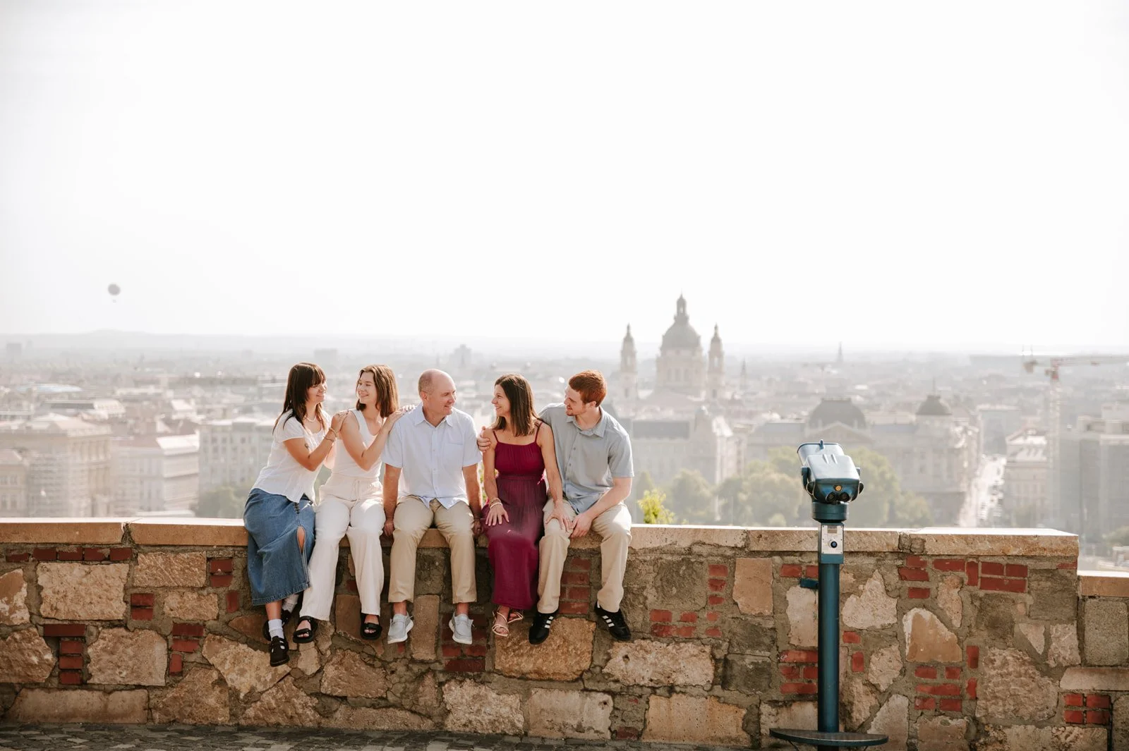 Five friends sitting on a stone wall overlooking a city skyline, with historic buildings and a prominent domed structure in the background, and a coin-operated viewing telescope nearby.
