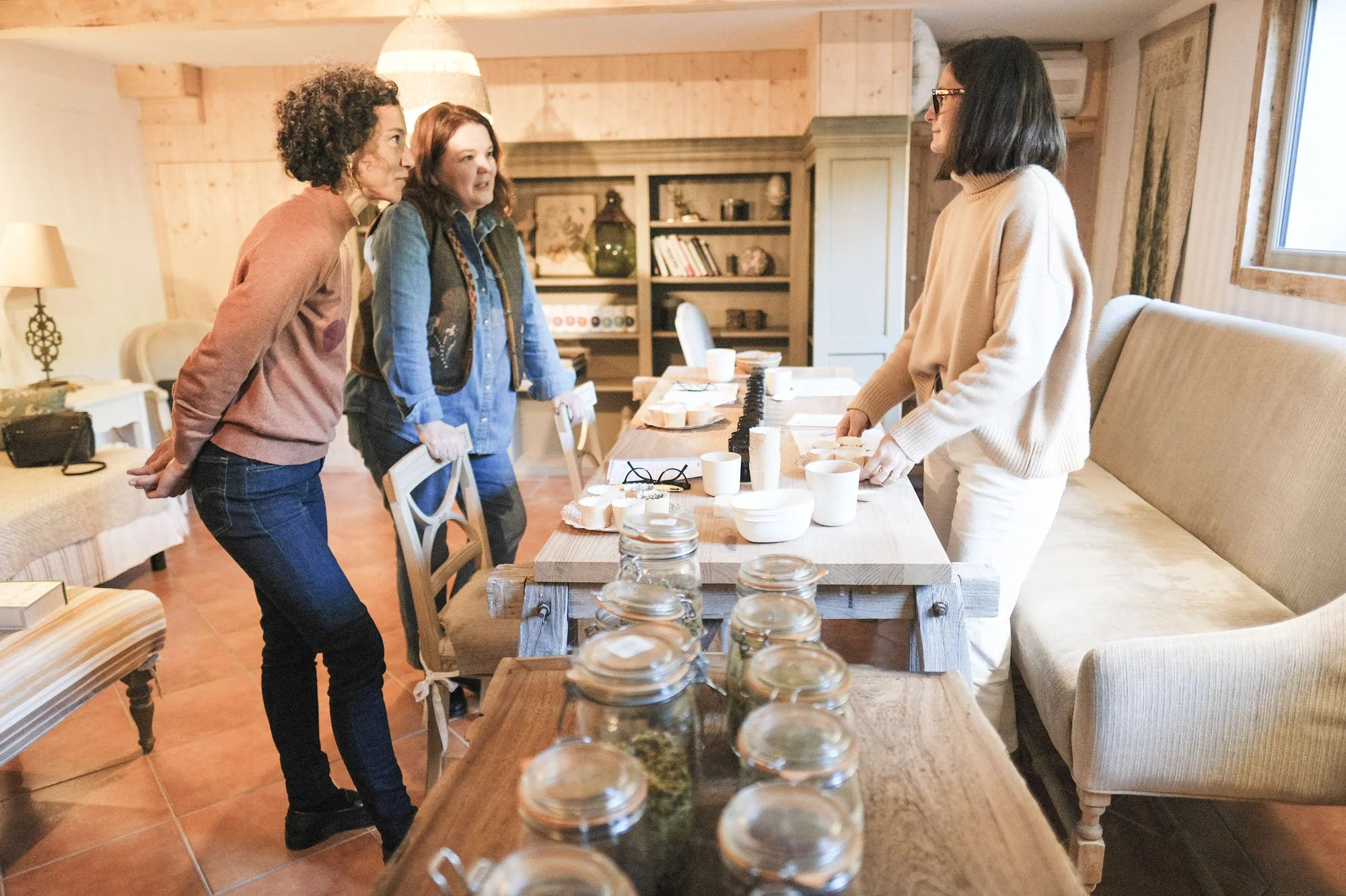 Trois femmes discutent autour d'une table en bois, à l'intérieur d'une pièce chaleureuse, avec des bocaux en verre au premier plan et des étagères de livres en arrière-plan.