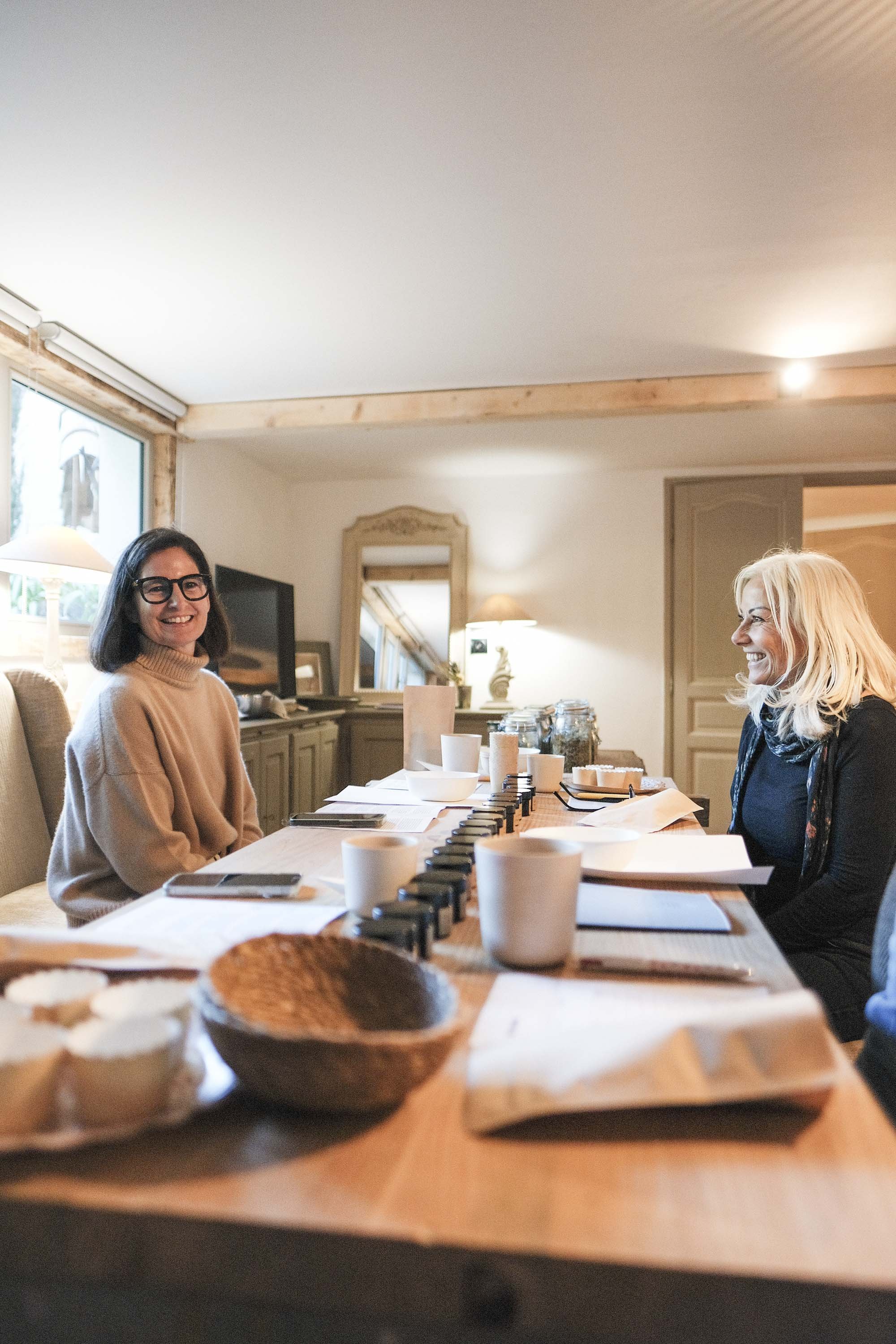 Deux femmes assises à une table en bois, souriant et discutant dans une pièce bien éclairée, avec des objets de cuisine et un miroir en arrière-plan.