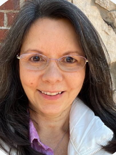 A woman with long dark hair, wearing glasses and a white jacket, smiling outdoors in front of a brick wall and rocks.