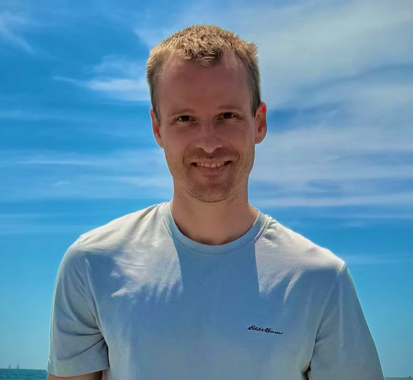A smiling man with short light hair wearing a white t-shirt, standing outdoors against a background of a blue sky with wispy clouds.