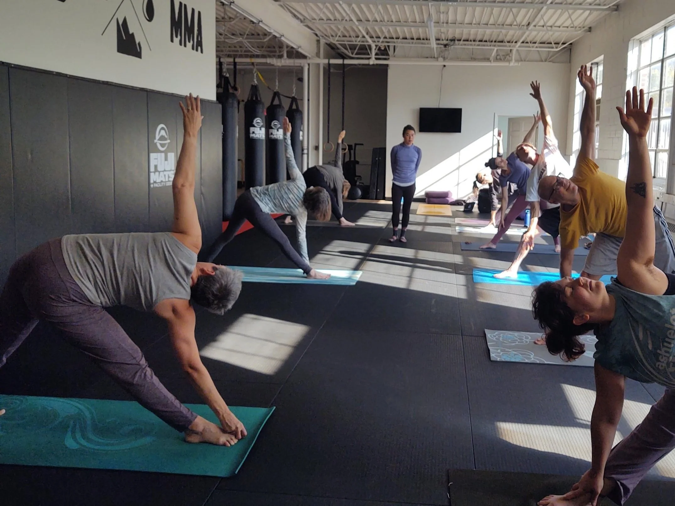People participating in a yoga class in a gym, performing side stretches while on yoga mats, with a instructor observing.