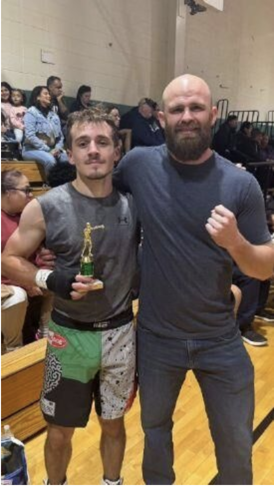 Ed, a young fighter from Elements MMA, stands proudly holding a trophy next to his coach, both smiling after a successful debut fight at Scrub Scraps 6. They pose inside a gymnasium with spectators in the background.