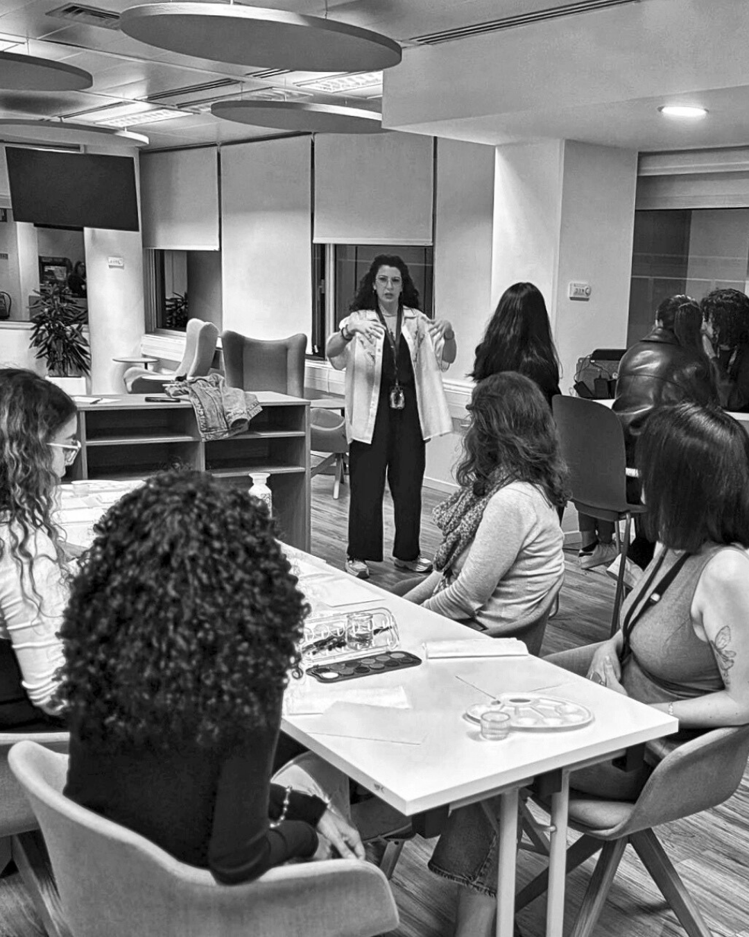 A woman giving a presentation in a conference room to a seated audience. The room has modern design elements and several tables with attendees.