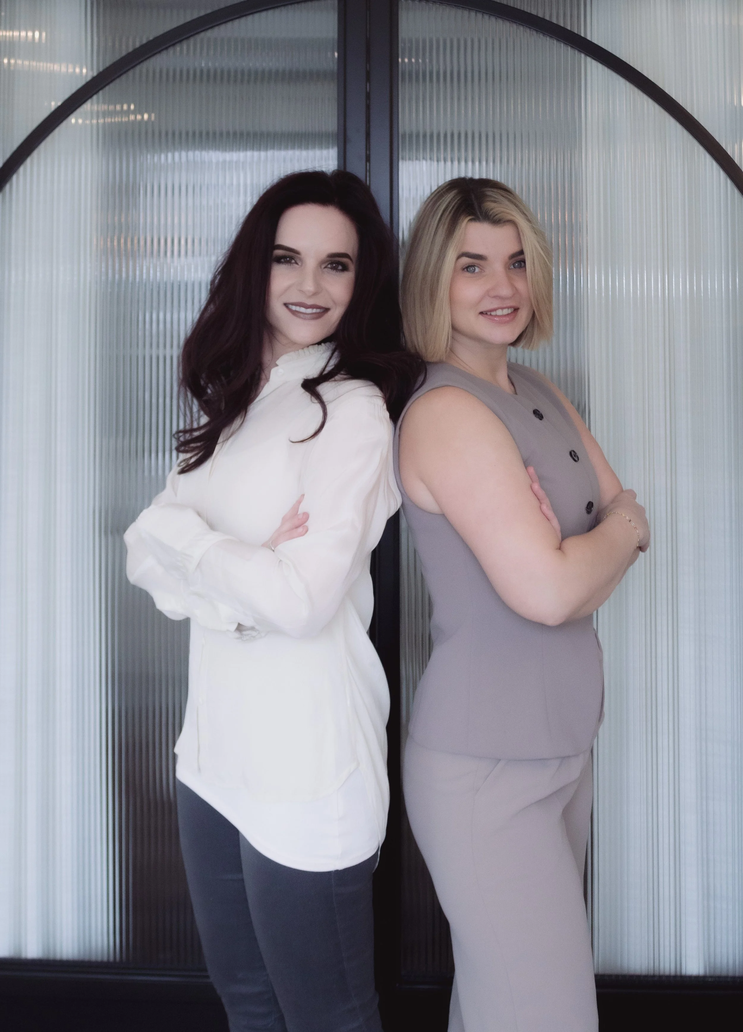 Two women standing back to back with arms crossed in front of a glass door, smiling.