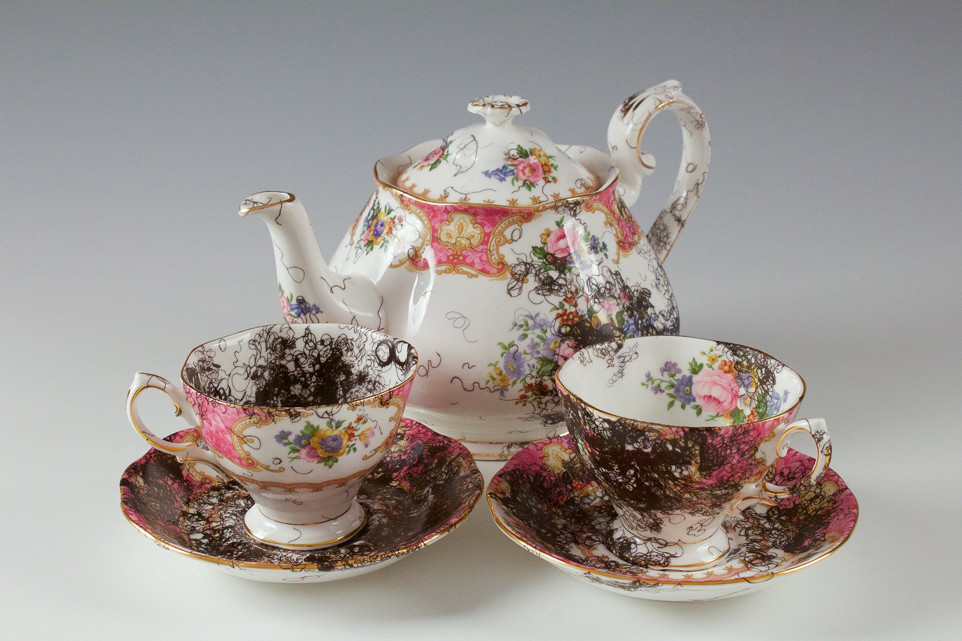 A vintage, china teapot and two cups and saucers with floral decorations is overlaid with decal drawings of curly, black hair.