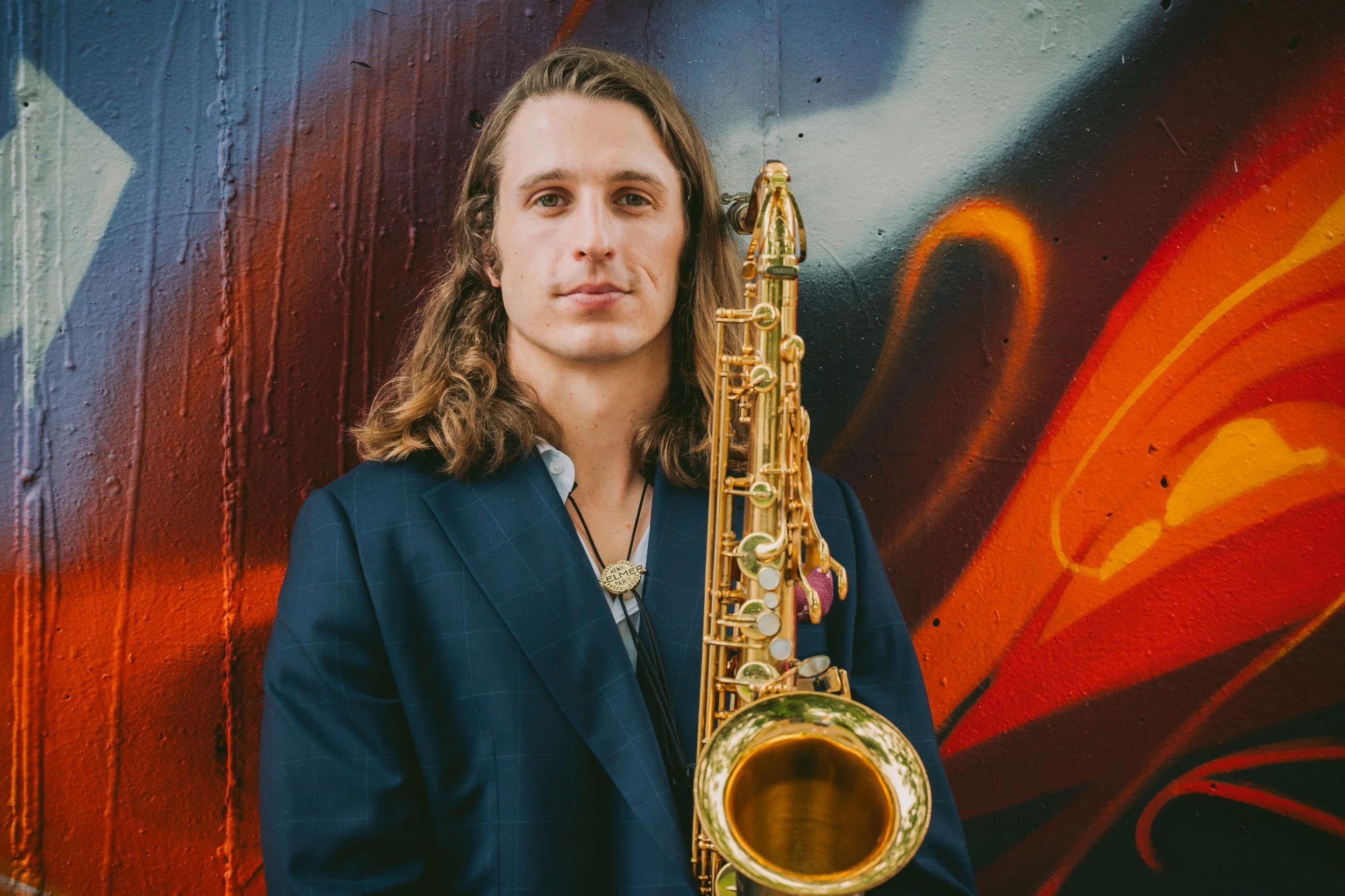 A man with long, wavy hair in a checkered blazer holding a gold alto saxophone, standing in front of a colorful graffiti wall.