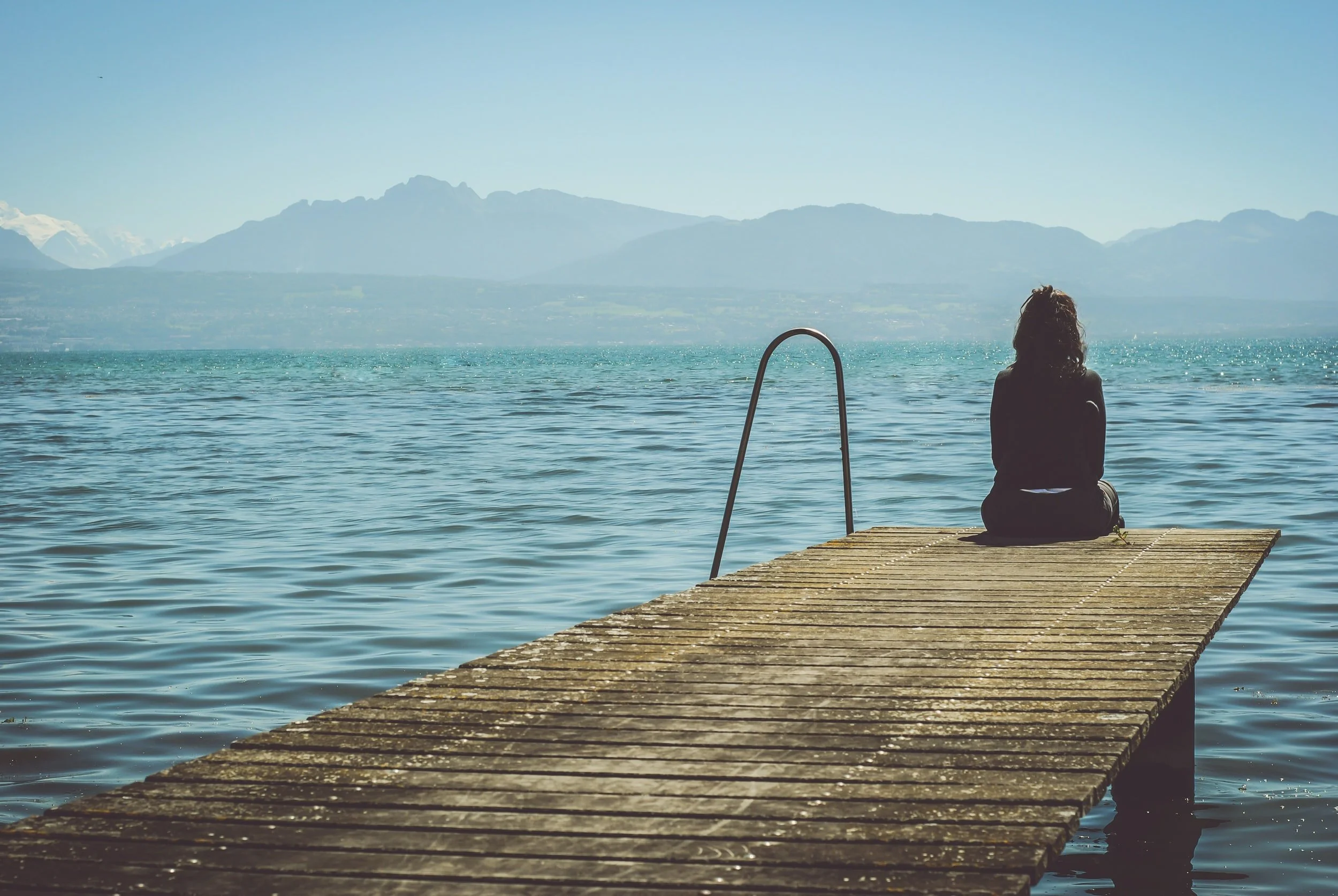 a woman sits on a dock overlooking a lake and mountains