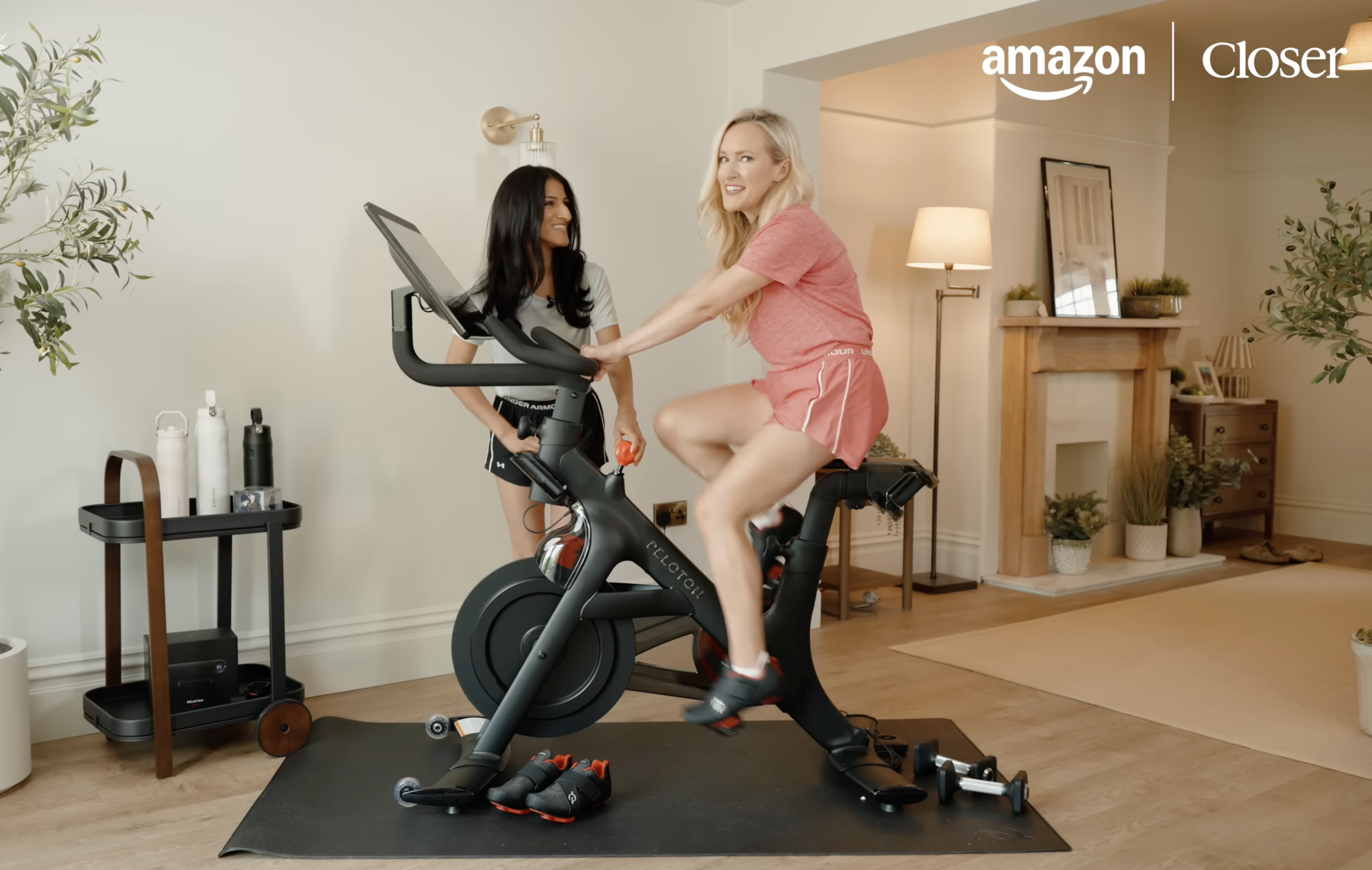 A blonde woman in pink workout clothes is riding a stationary bike indoors, while a brunette woman in a gray shirt and shorts stands beside her, smiling. The room has a cozy, modern decor with a fireplace, mirror, potted plants, and a small table with water bottles. The background includes a cream-colored wall and warm lighting.