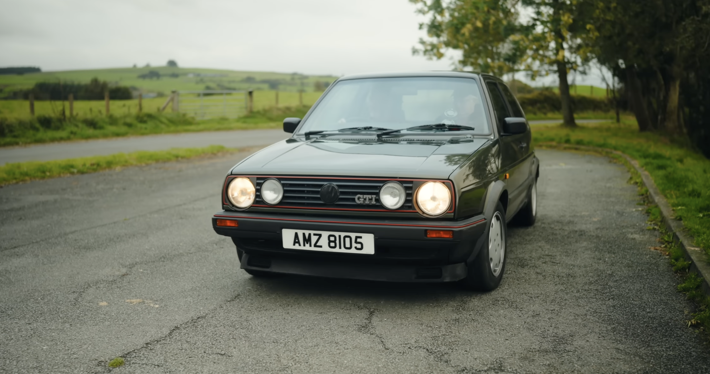 A black Volkswagen Golf GTI parked on a rural road with green fields, trees, and overcast sky in the background.