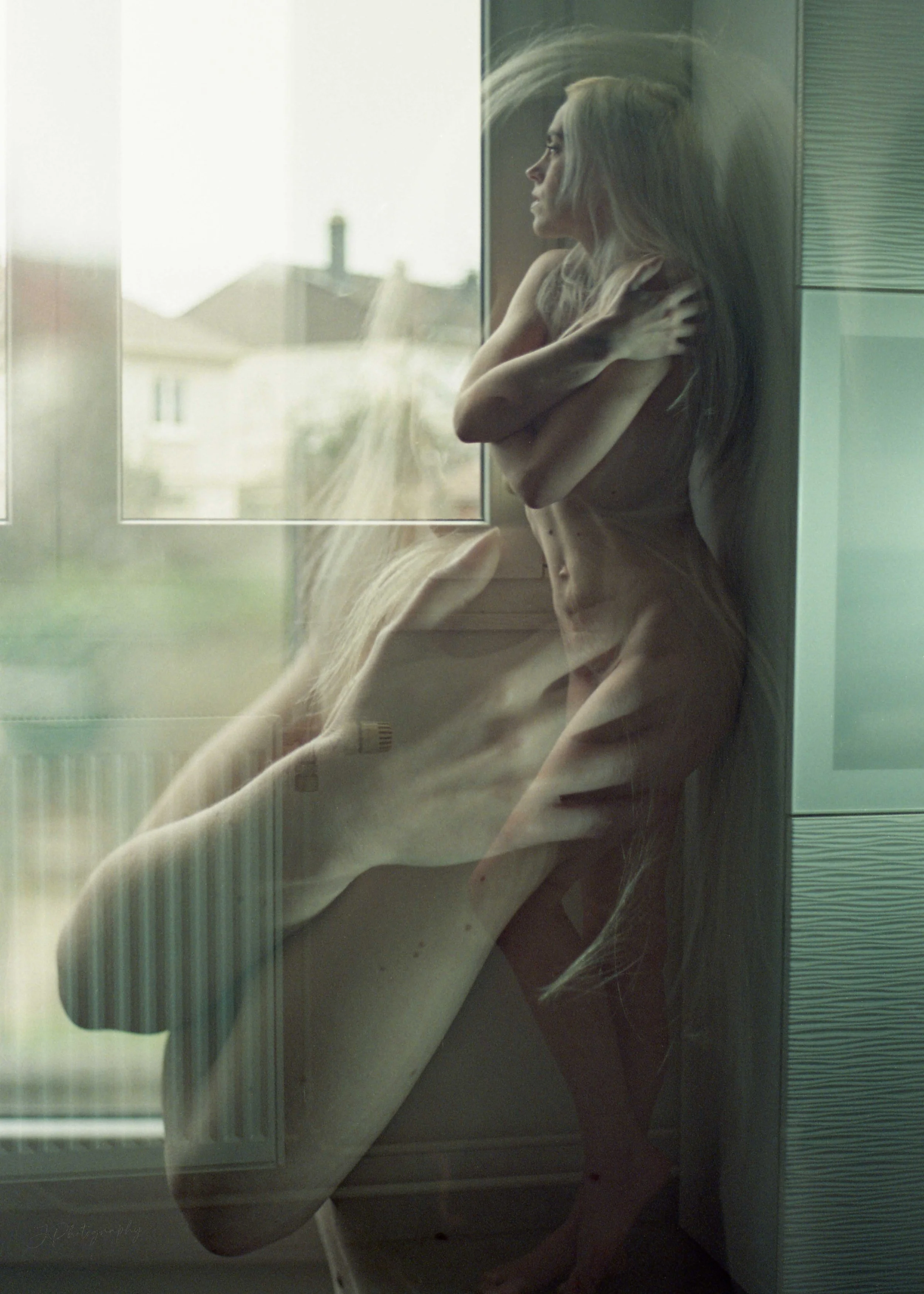 Une femme nue debout près d'une fenêtre, regardant à l'extérieur, avec une double exposition qui montre une autre silhouette féminine assise à l'intérieur.