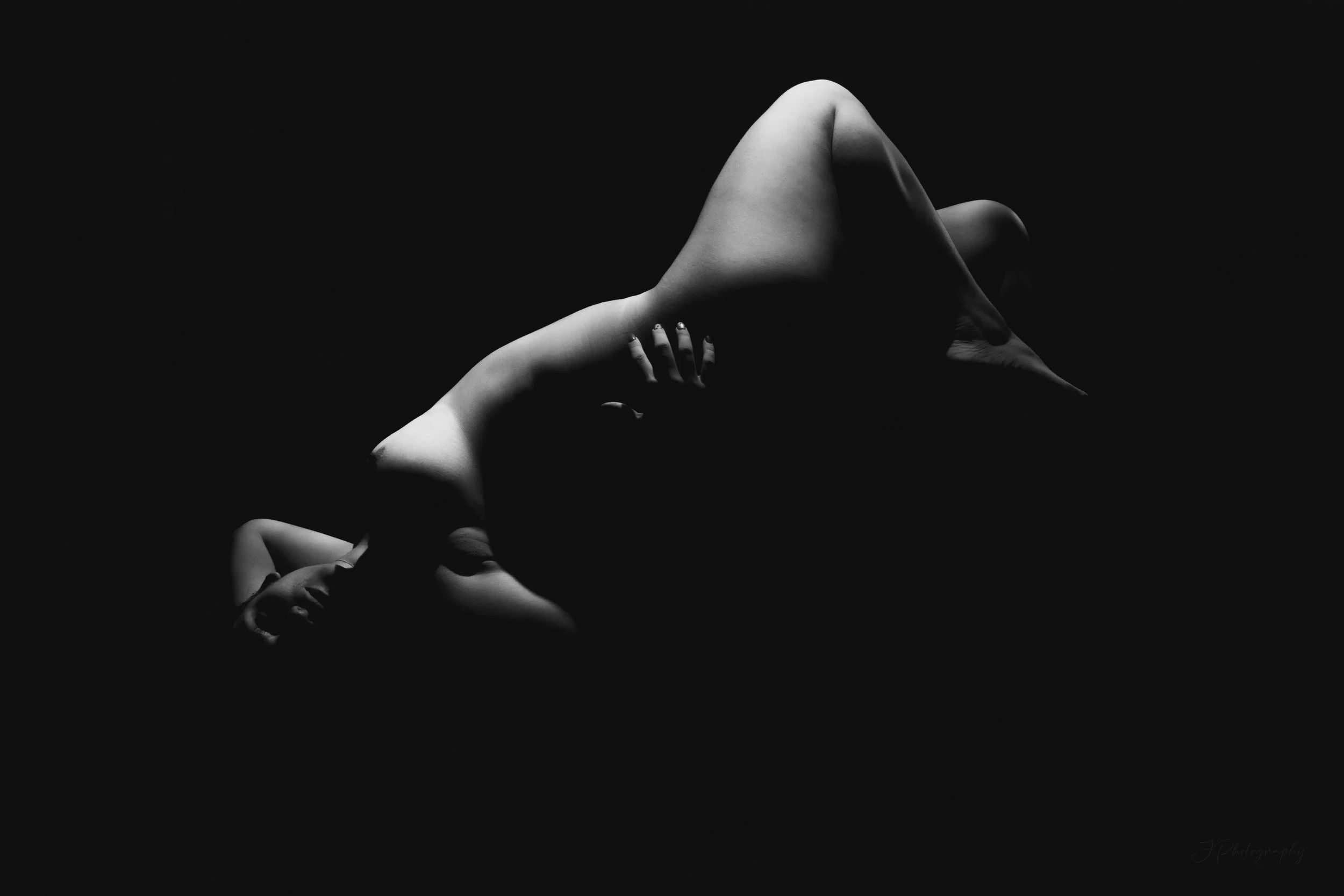 Photo en noir et blanc d'une femme nue allongée, vue de profil, le visage dans l'ombre, dans une position flottante ou de lévitation, avec une lumière douce mettant en valeur ses formes et tatouages. Technique de lévitation.