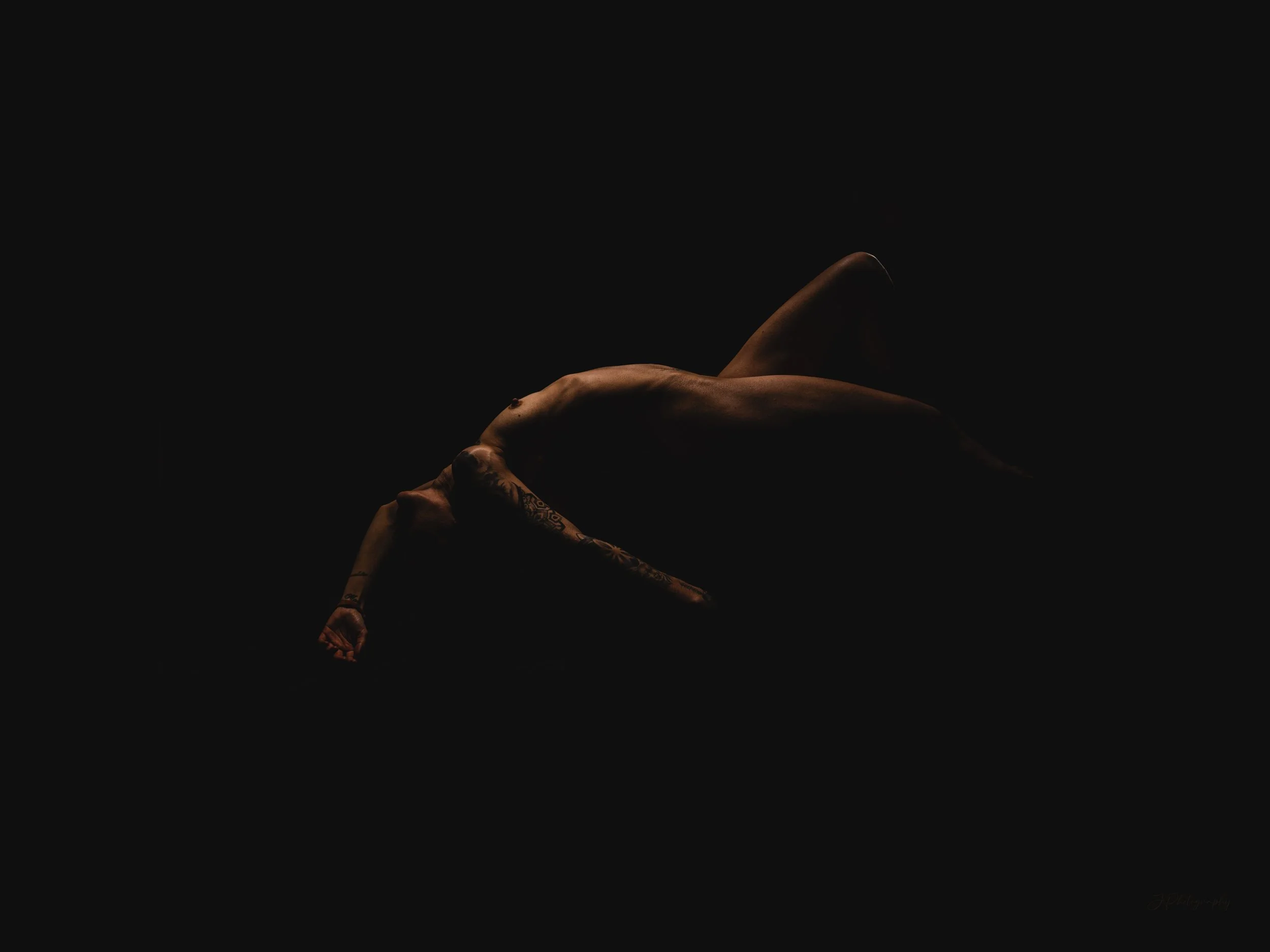 Une femme nue repose sur le côté dans l'obscurité, éclairée partiellement, mettant en valeur sa silhouette et ses tatouages.