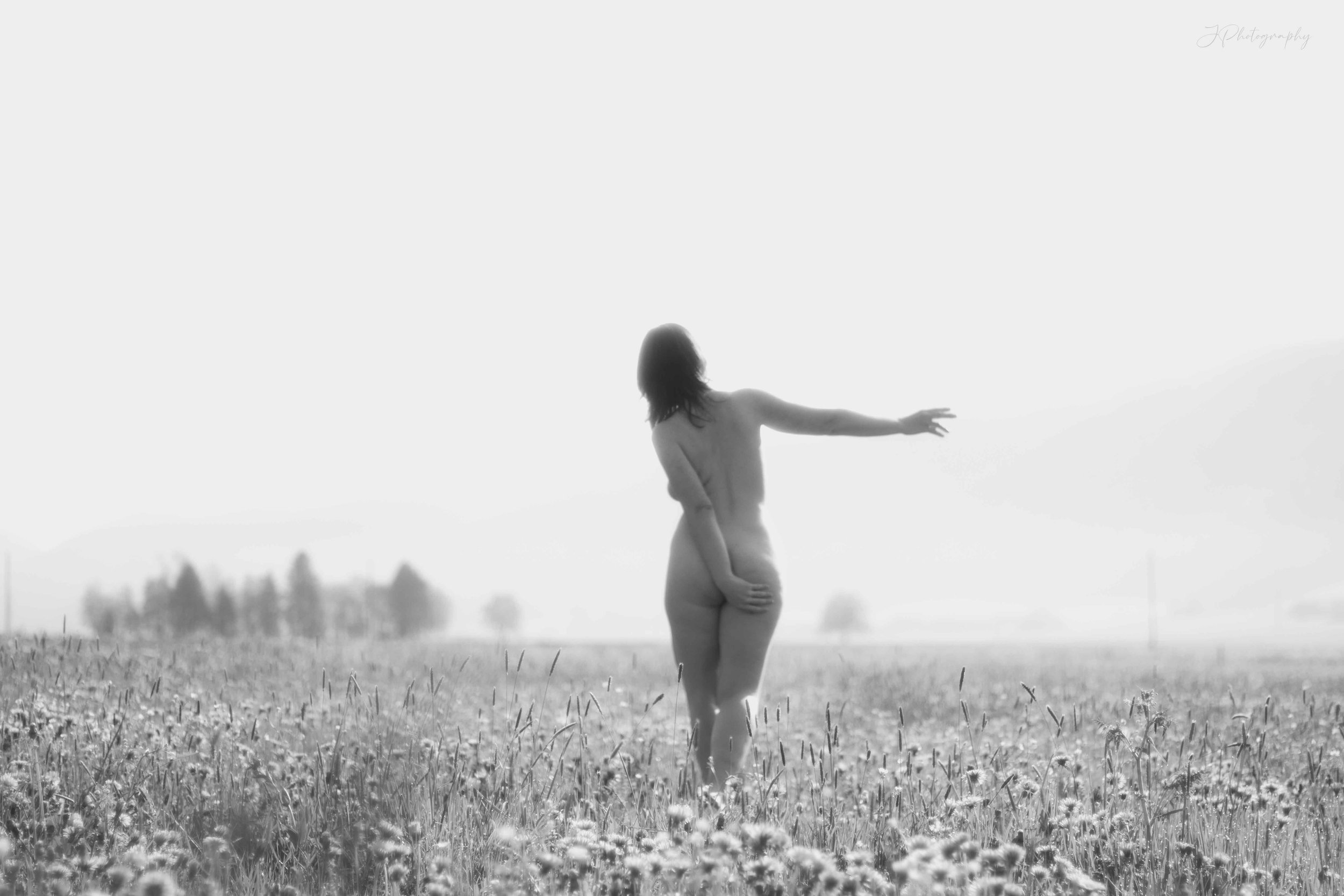Une femme nue dans un champ, en train de faire un geste avec son bras droit. La photo est en noir et blanc, avec un paysage de nature en arrière-plan.