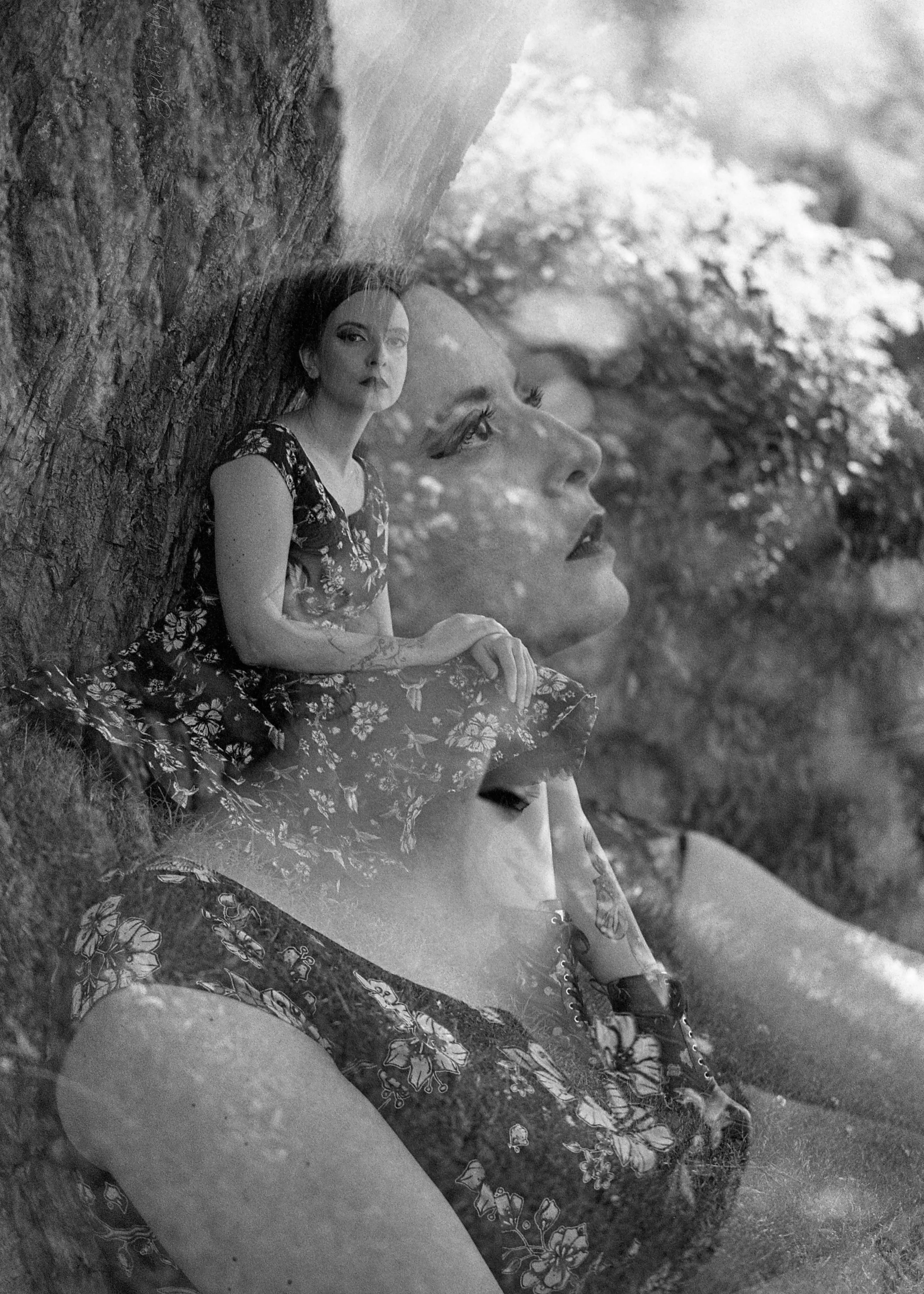 Une photographie en noir et blanc représentant deux femmes, une jeune assise contre un arbre et une autre grande en arrière-plan avec un visage en gros plan. La scène est artistique et rêveuse.