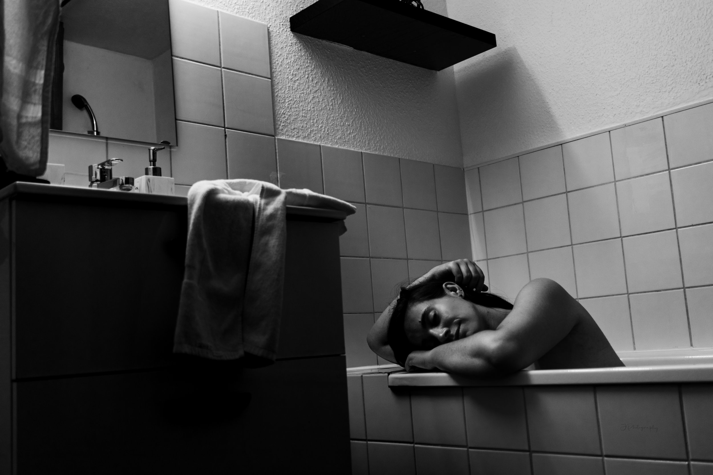 Une femme repose dans une baignoire en regardant vers le haut, dans une salle de bain en noir et blanc avec un lavabo à gauche.