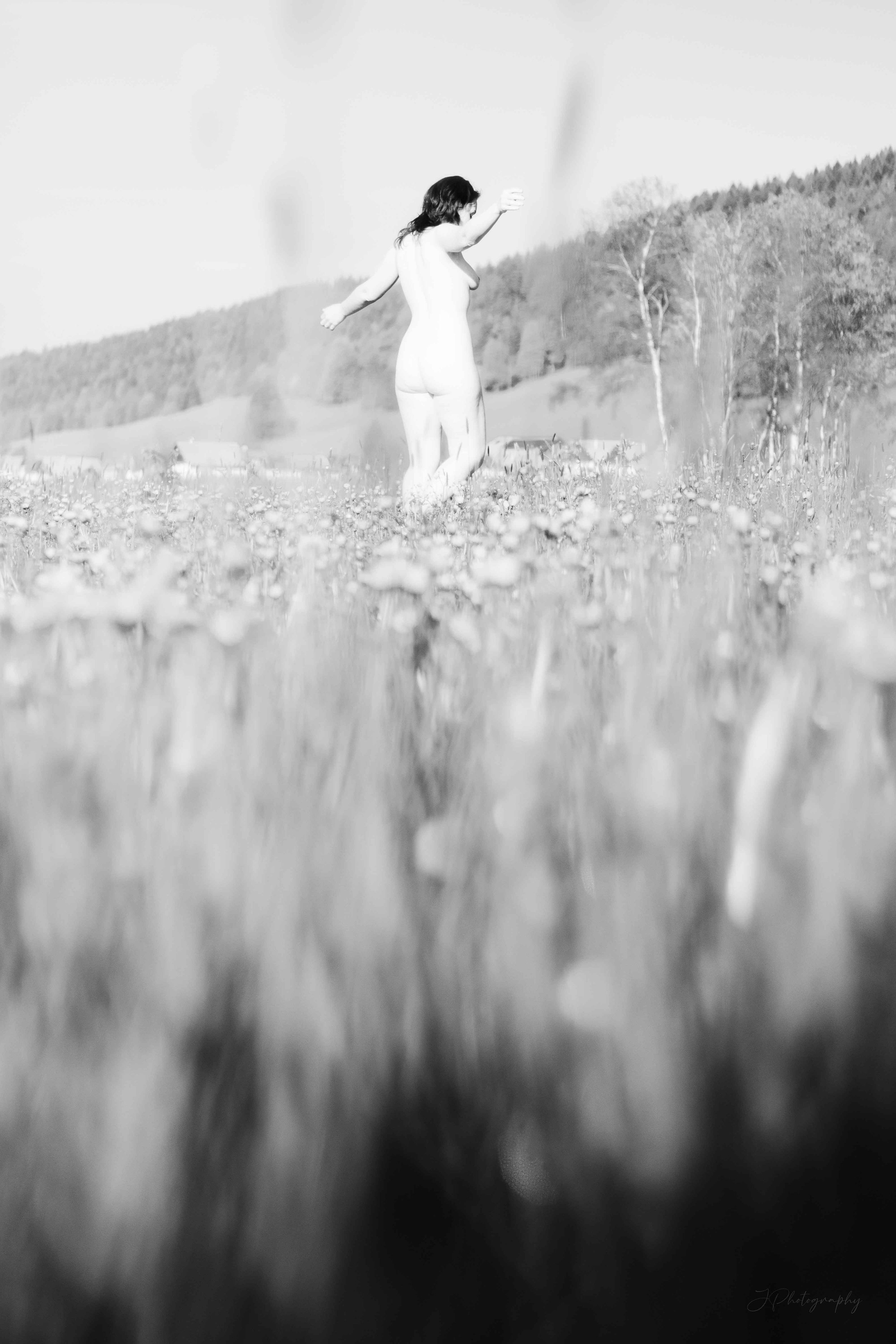 Une femme nue marche dans un champ avec des collines boisées en arrière-plan, vue de côté, en noir et blanc.