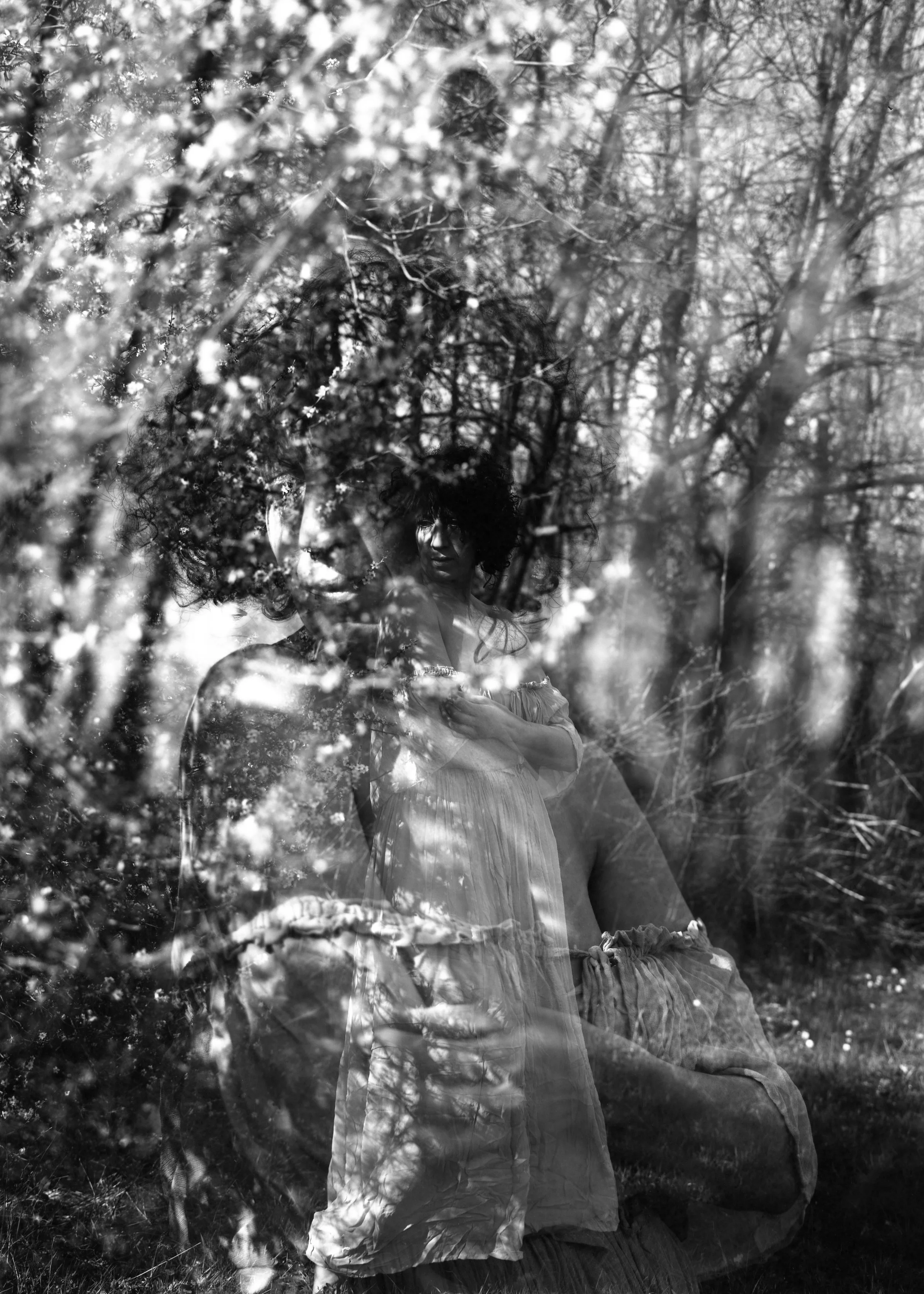 Une femme est photographiée en noir et blanc, avec un reflet ou une superposition d'arbres et de fleurs. Elle porte une robe légère et regarde vers l'avant.