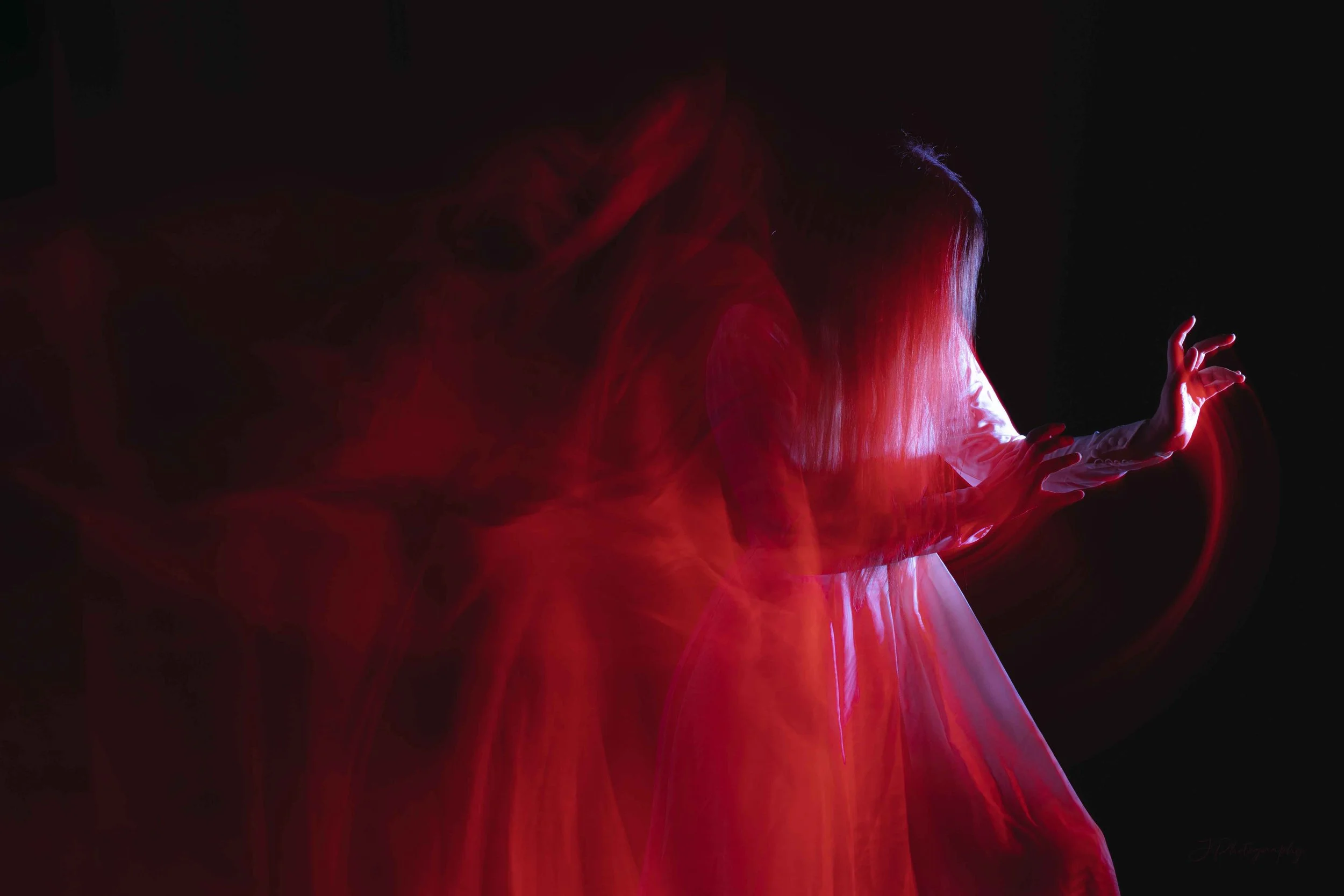 Une femme avec une robe blanche et un long manteau rouge, dont la tête est inclinée en avant, dans un environnement sombre. Des effets de lumière créent un mouvement dynamique autour d'elle.