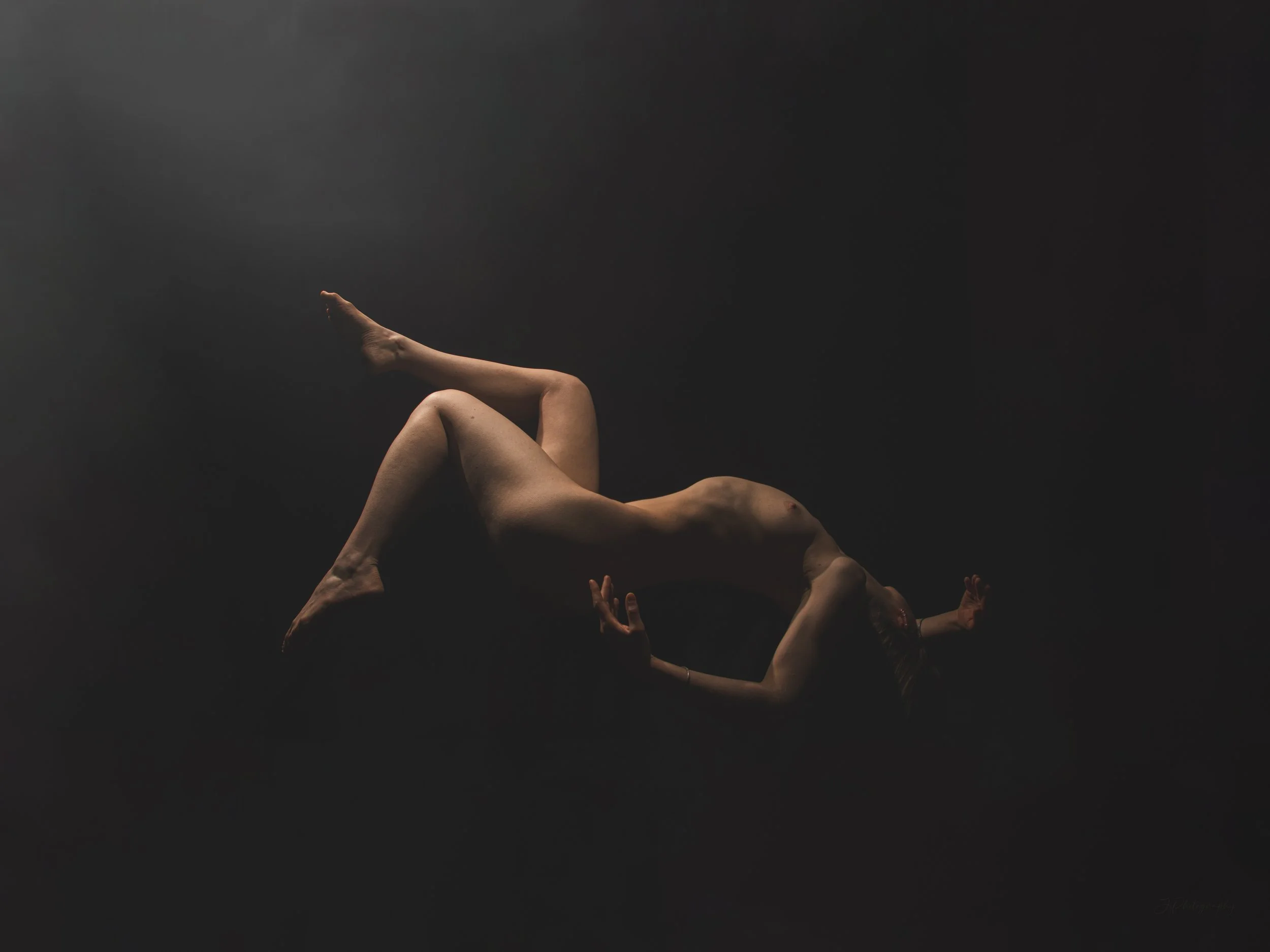 Une personne nue en position de flexion arrière sur un fond noir, avec un seul point de lumière mettant en valeur son corps.